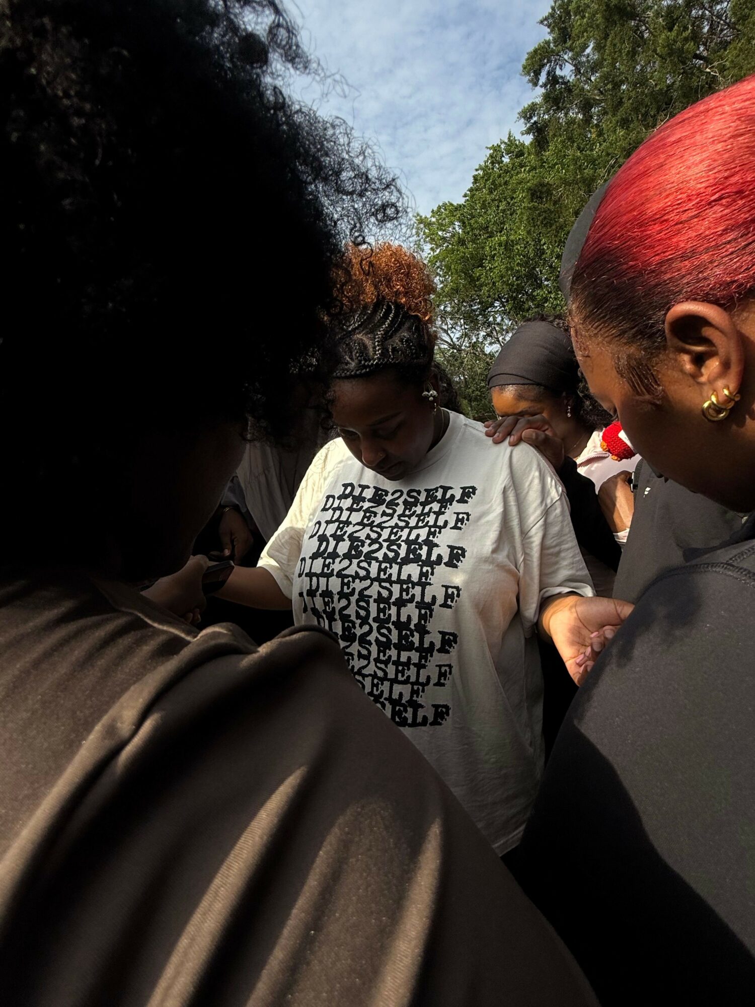 Group of people outdoors, some wearing white shirts with black text, with trees and sky in background.