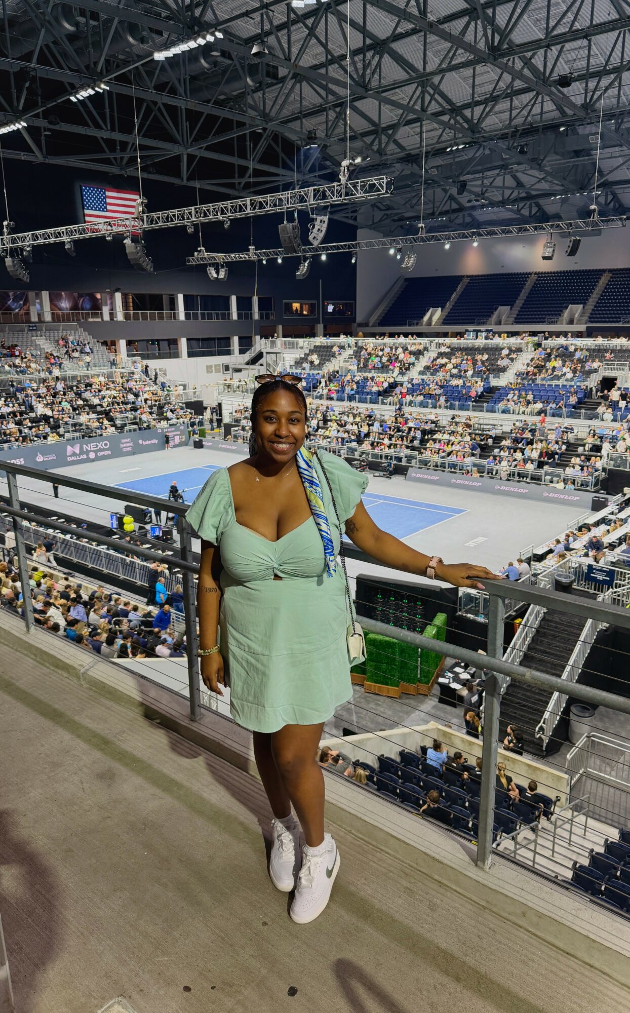 Woman standing in an indoor sports arena, smiling, with seating and a tennis court in the background.