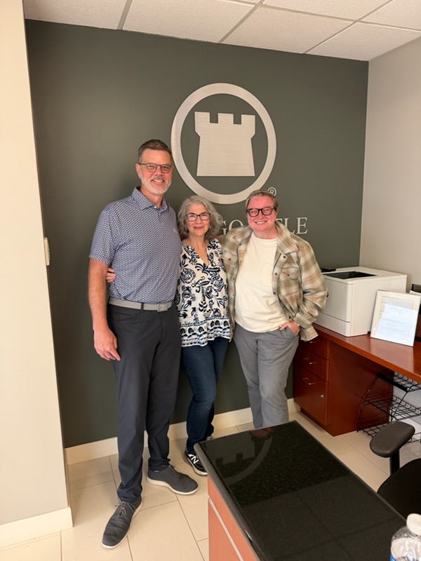 Three people standing indoors in front of a wall with a castle logo, smiling, with office furniture nearby.