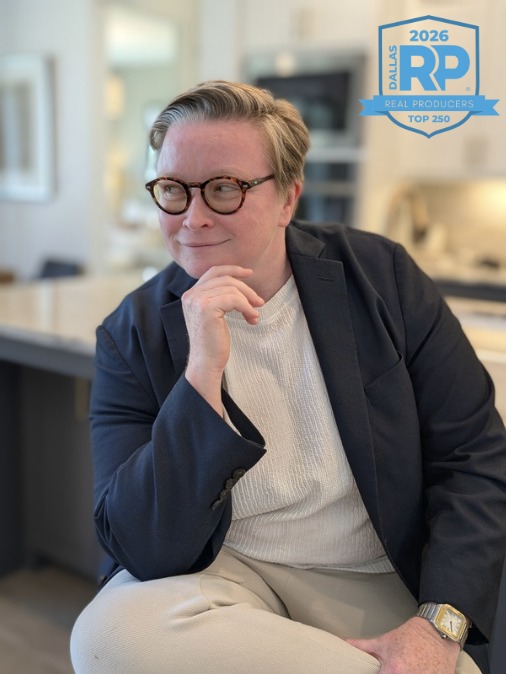 Person with short hair and glasses, sitting thoughtfully in a modern office, wearing a dark blazer and white shirt.