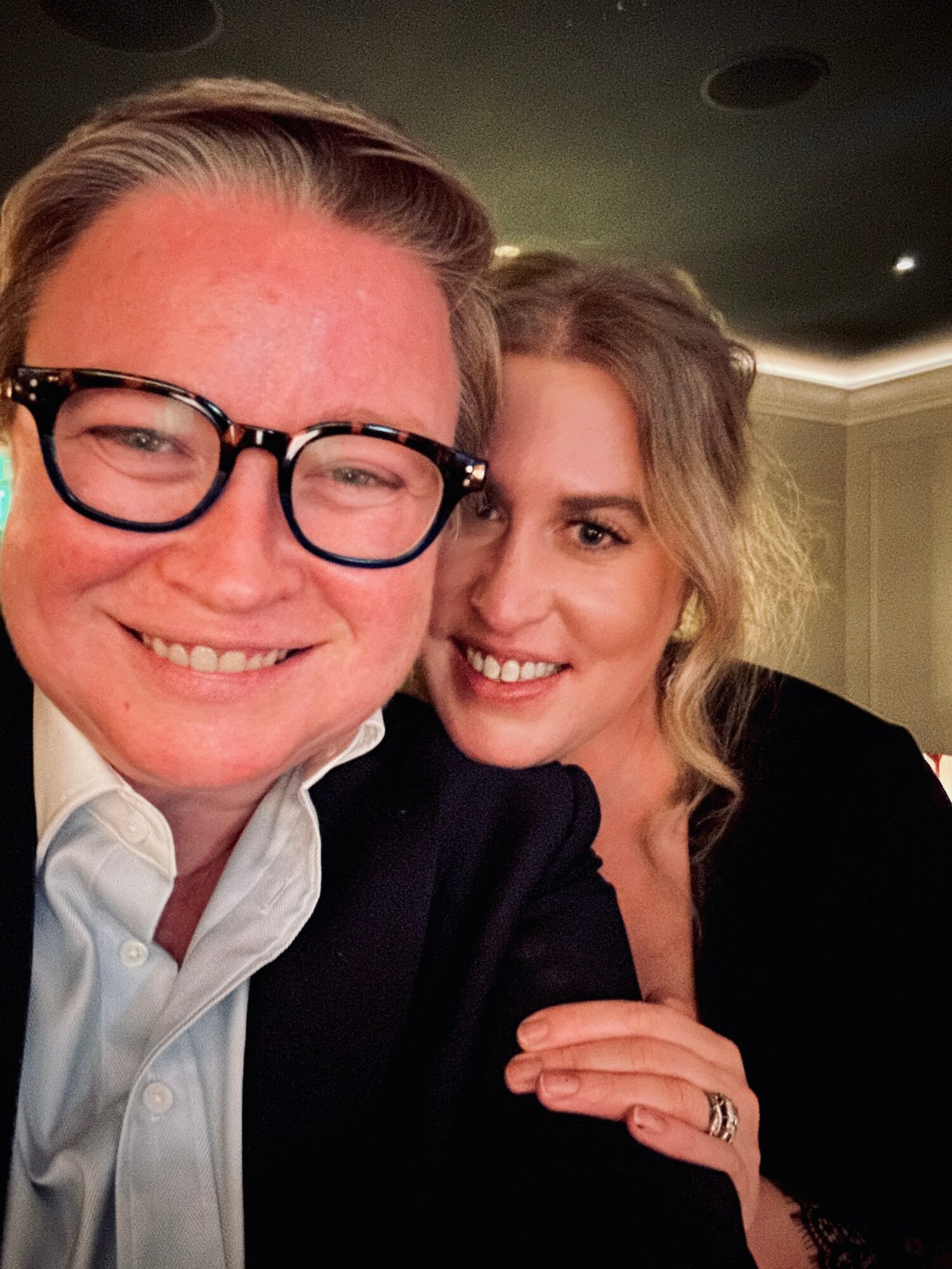 Two people smiling closely, one wearing glasses, in an indoor setting with warm lighting.