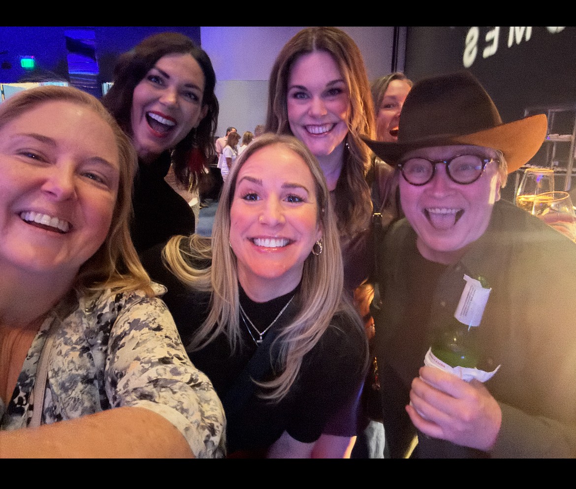 Group of five women smiling and taking a selfie at an indoor event, one wearing a hat and glasses, holding a drink.
