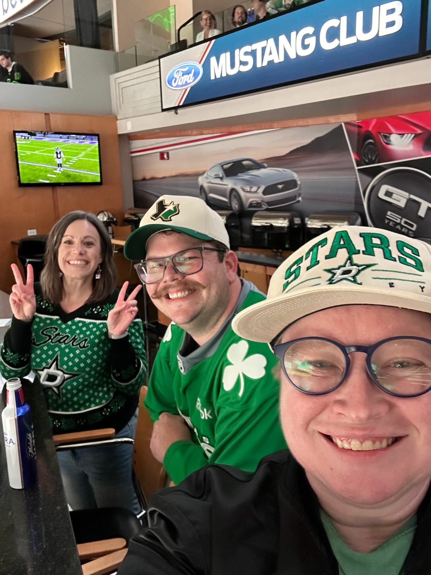 Two smiling people wearing green sports jerseys and glasses, making peace signs, in a sports bar with a TV showing a game.