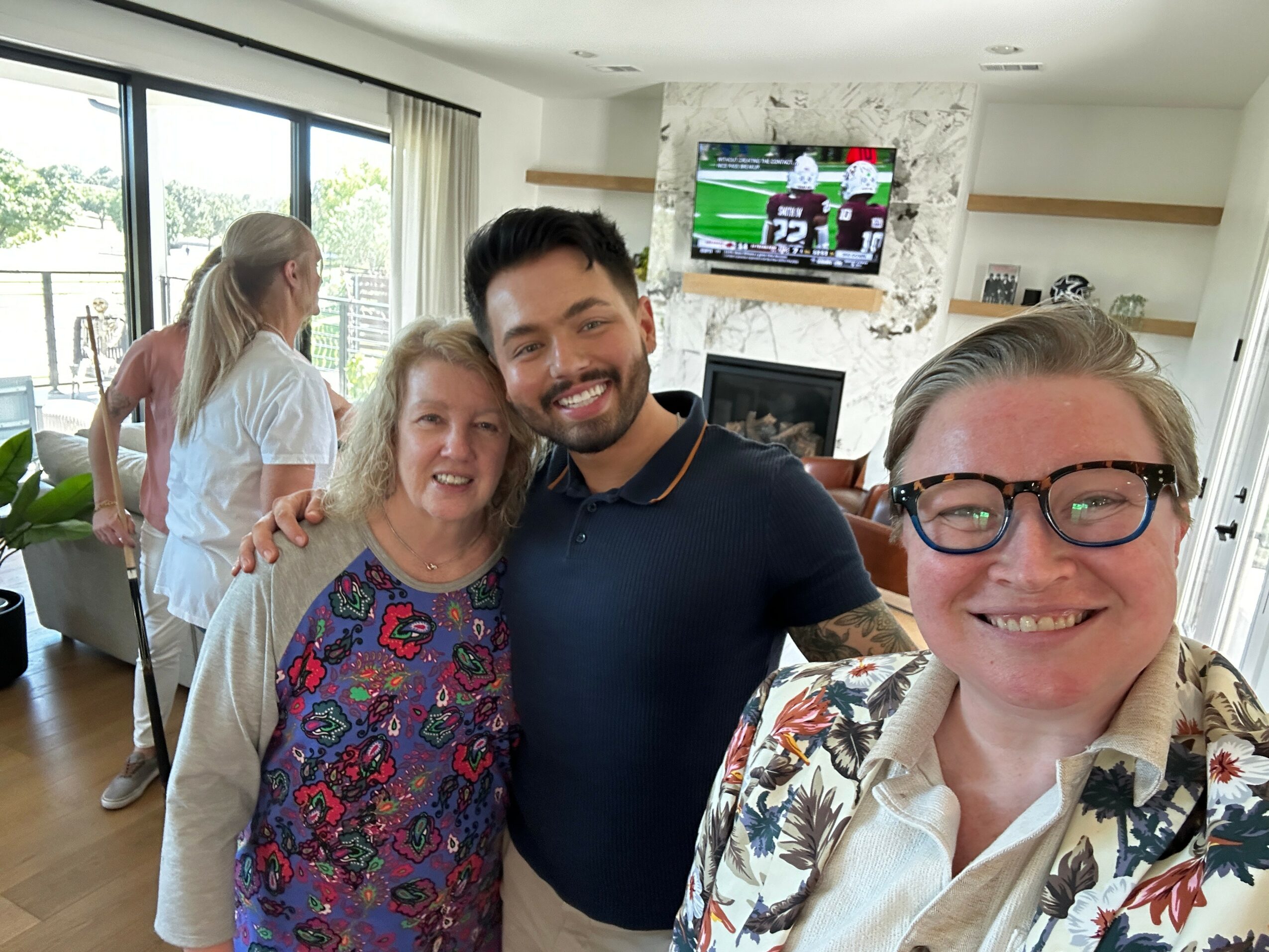 Four people smiling in a living room, with a TV showing a sports game in the background.
