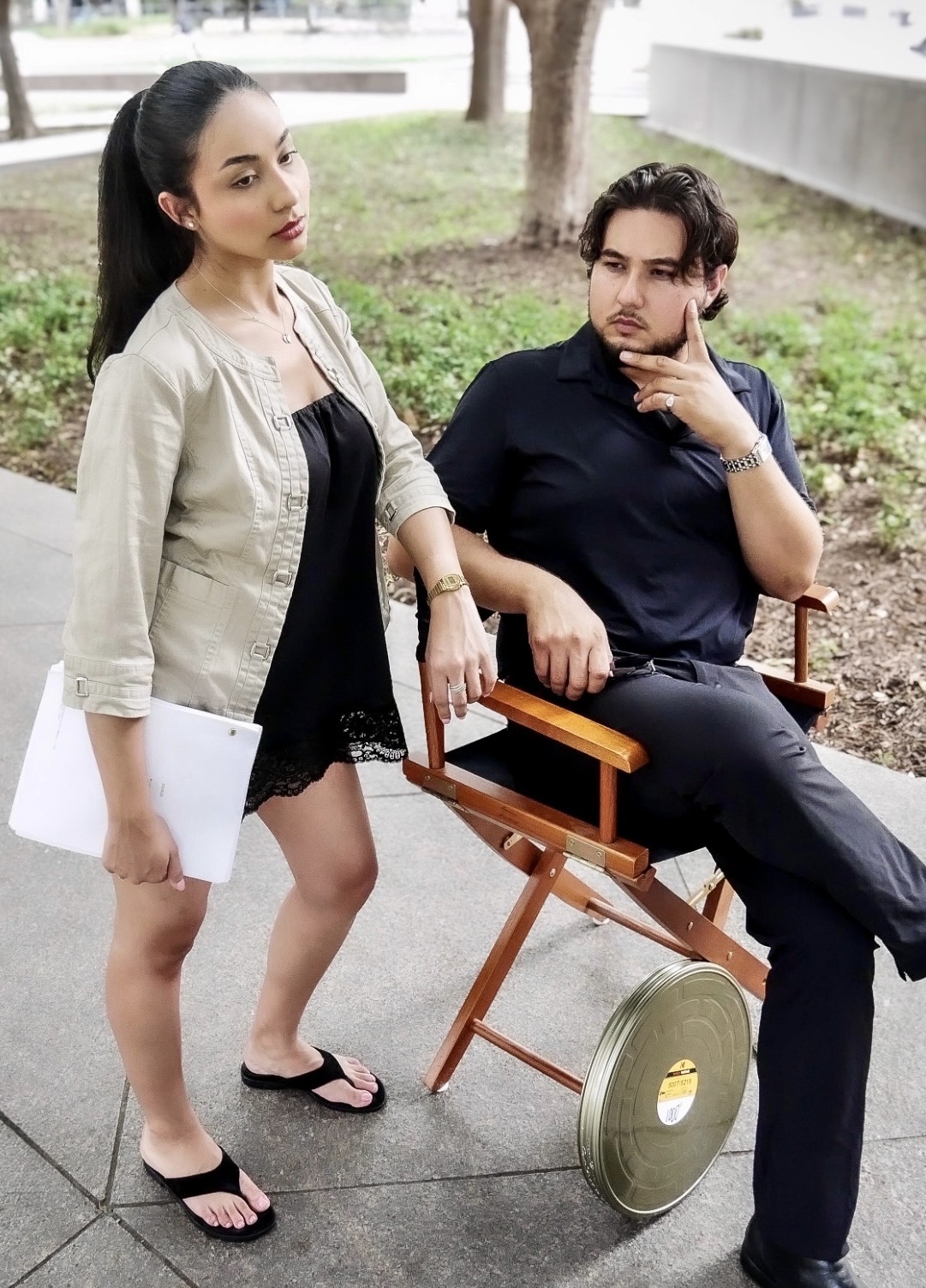 A woman standing outdoors with a man sitting on a wooden chair, holding a paper, in a park-like setting.