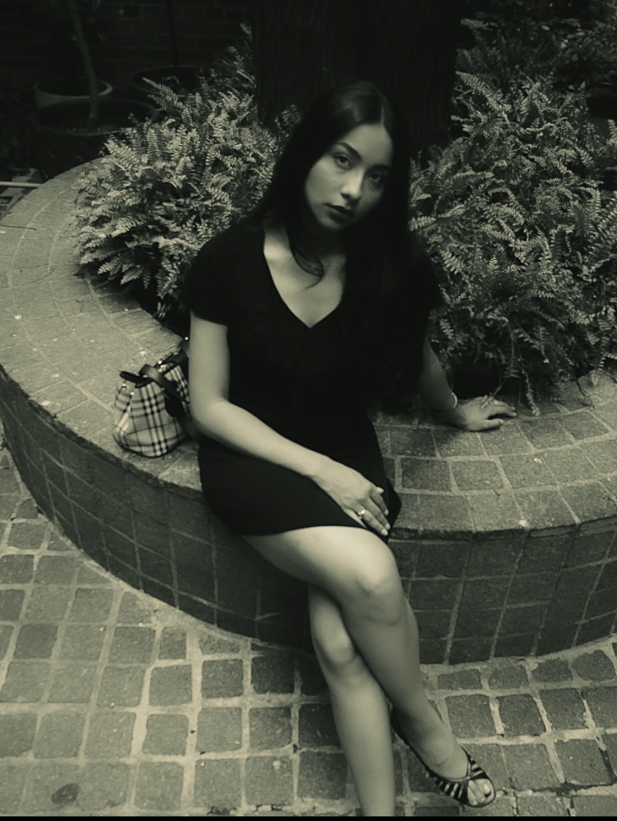 Young woman sitting on a curved brick wall with plants behind her, wearing a black dress and high heels.