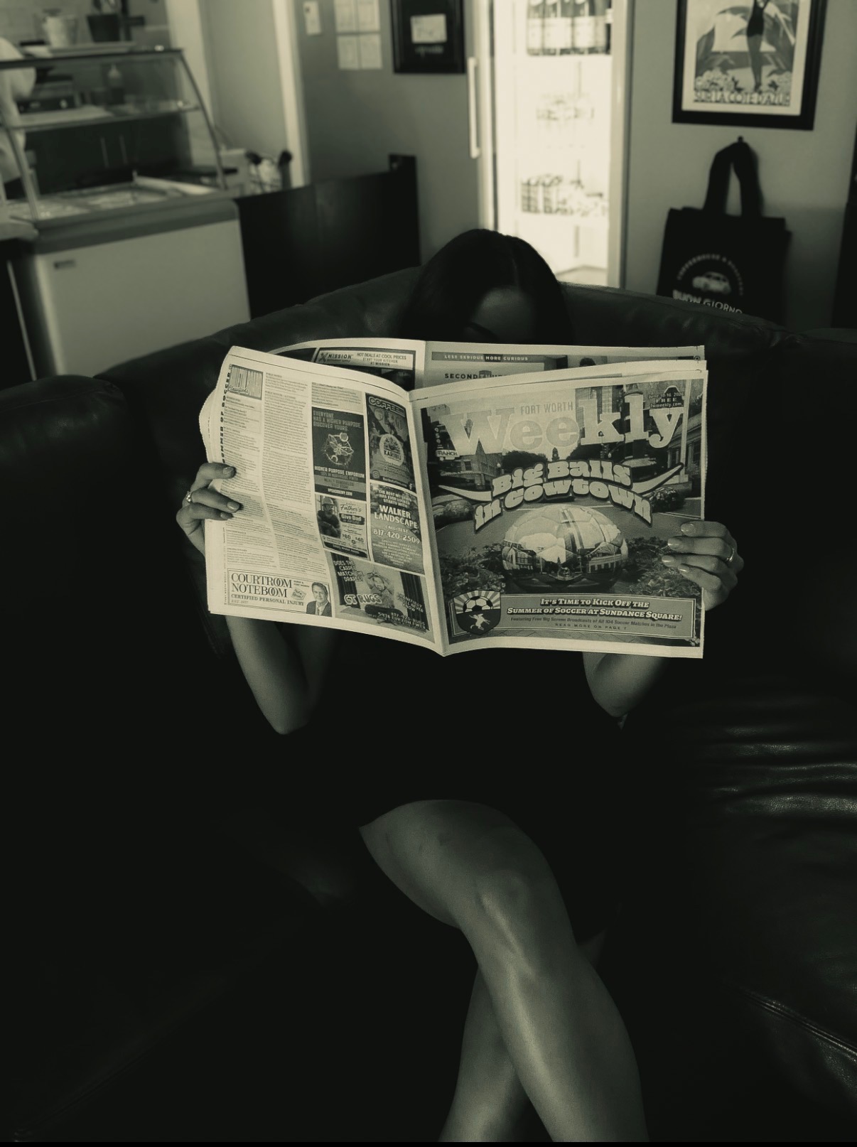 Person sitting on a couch reading a newspaper, with a kitchen and framed picture in the background.