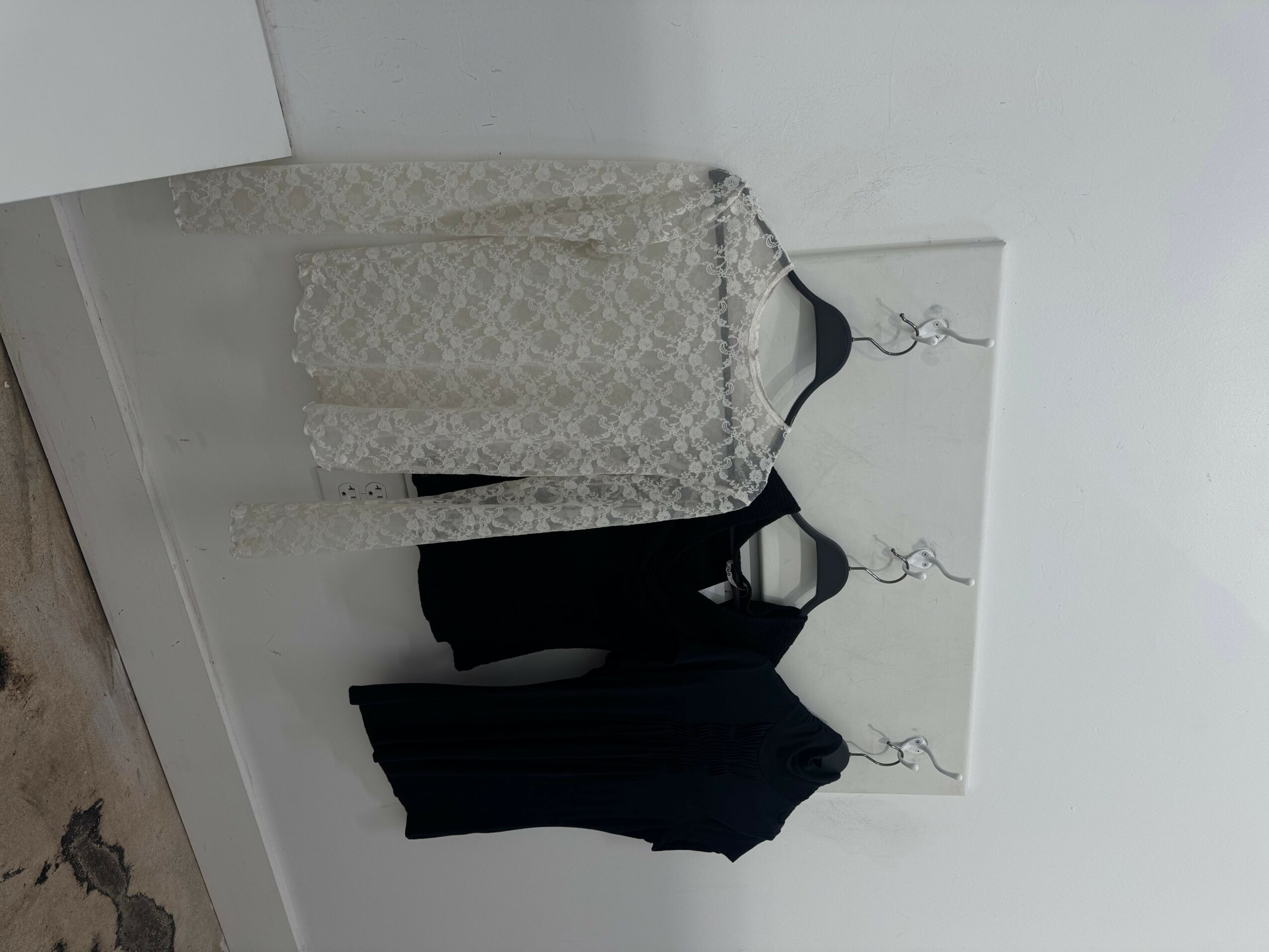 Clothes hanging on a wall, including a black dress, a black top, and a white patterned dress, with a mirror behind them.