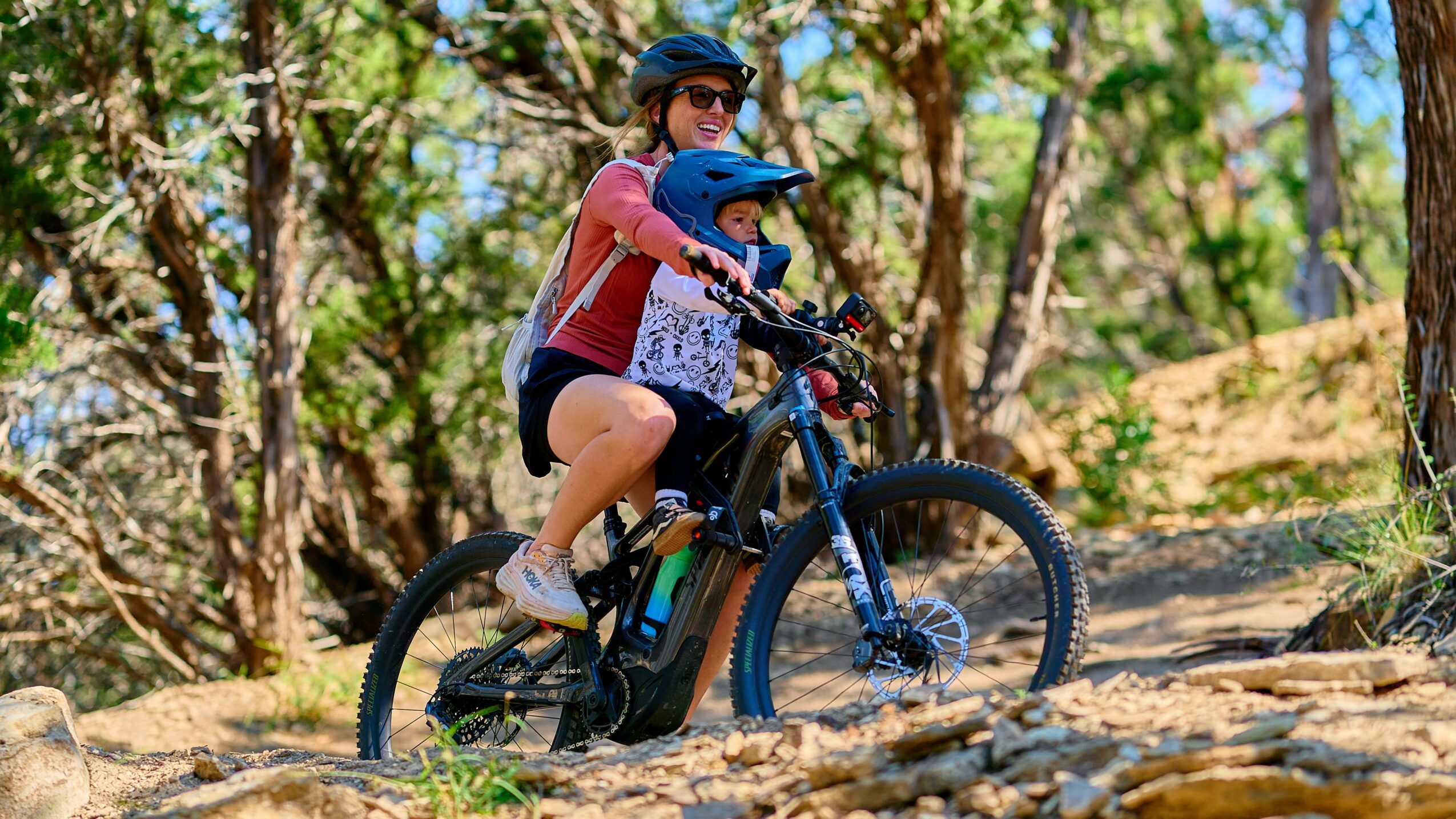 Two children riding a mountain bike on a trail surrounded by trees and sunlight, wearing helmets and casual clothing.