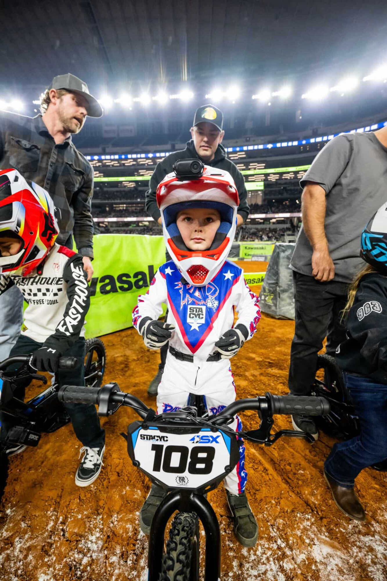 Young motocross rider in gear and helmet, standing with bike on dirt track, surrounded by adults, stadium lights overhead.