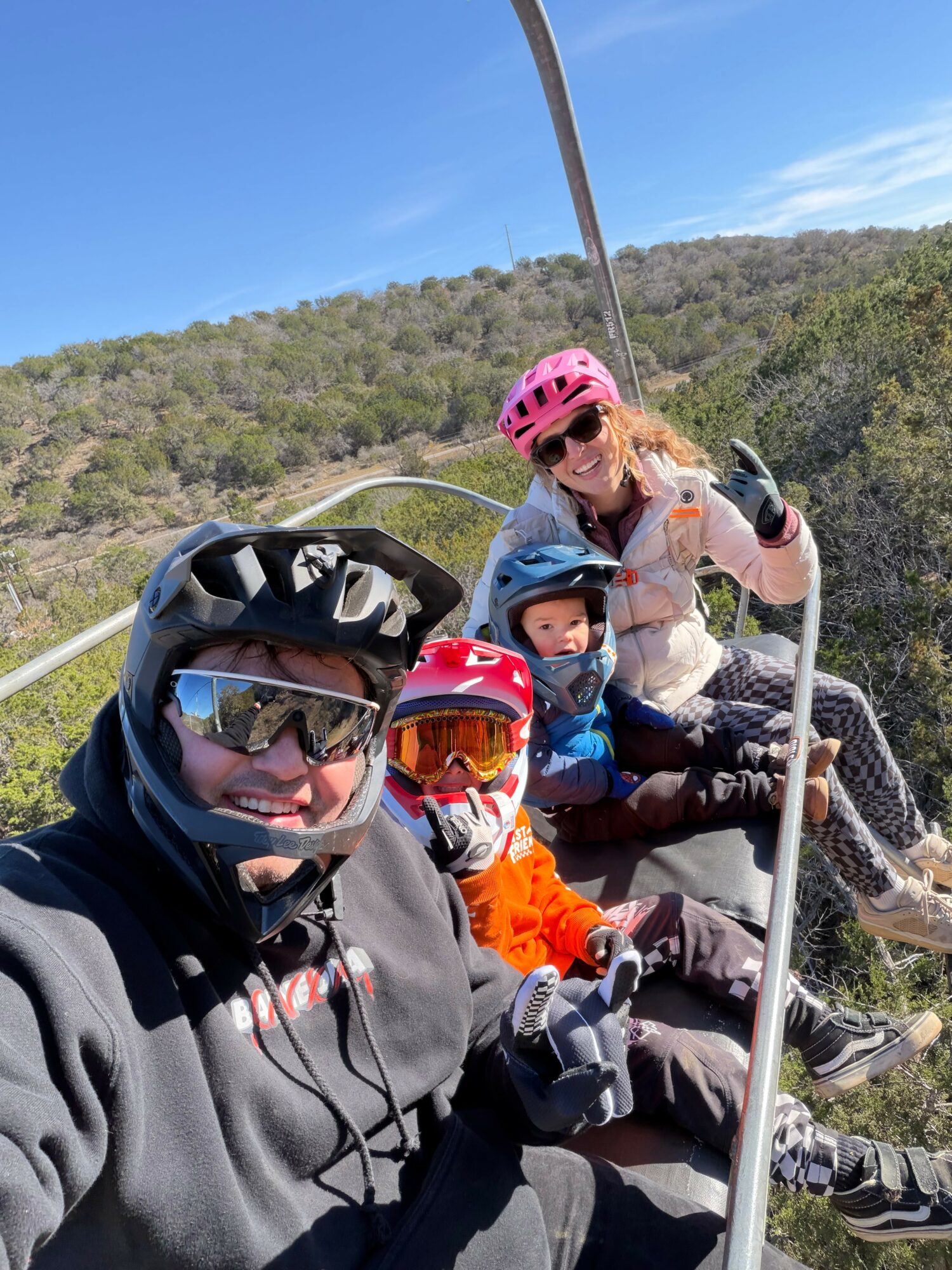 Four people wearing helmets and goggles sit on a ski lift, smiling and waving, with a hilly landscape and blue sky in the background.