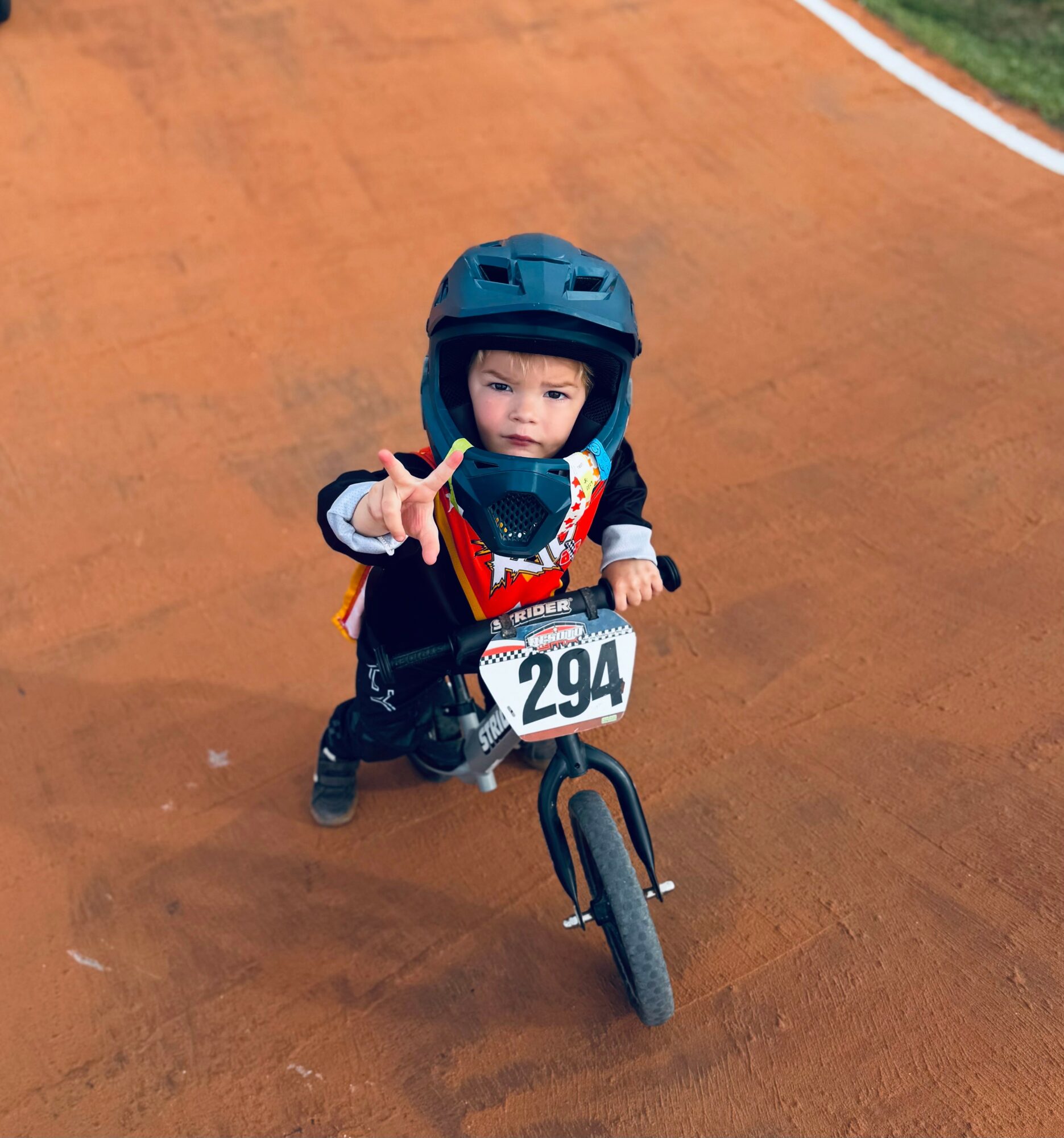 Young child riding a balance bike on a track, wearing a helmet and safety vest, reaching out with one hand.
