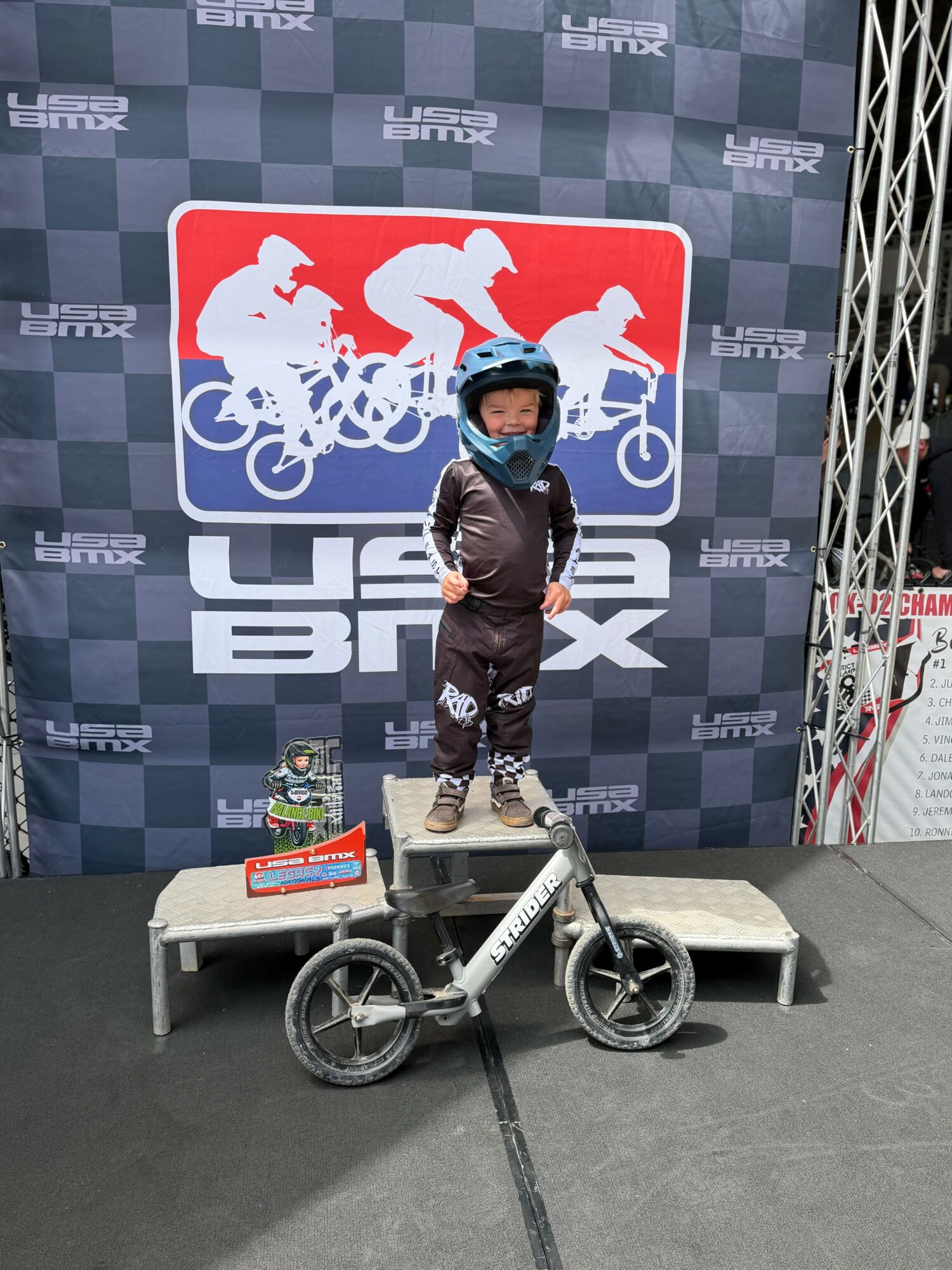 Child wearing a helmet standing on a platform with a balance bike, in front of a US BMX backdrop.