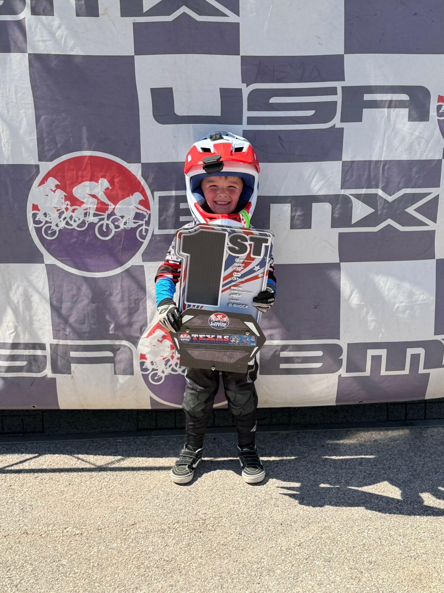 Child in racing gear holding a trophy and a bag, standing in front of a USA-themed backdrop.
