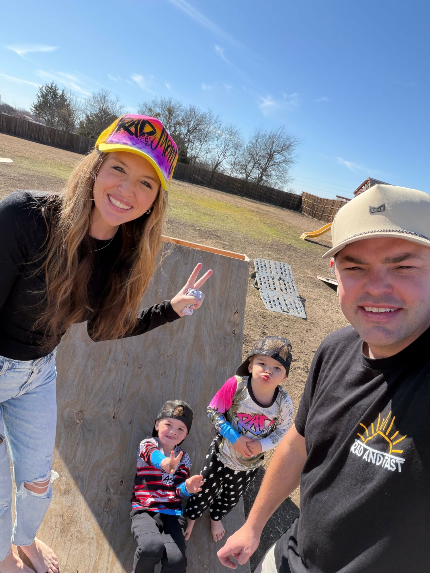 Four people outdoors, two children and two adults, smiling and making peace signs, with a skate ramp and trees in background.