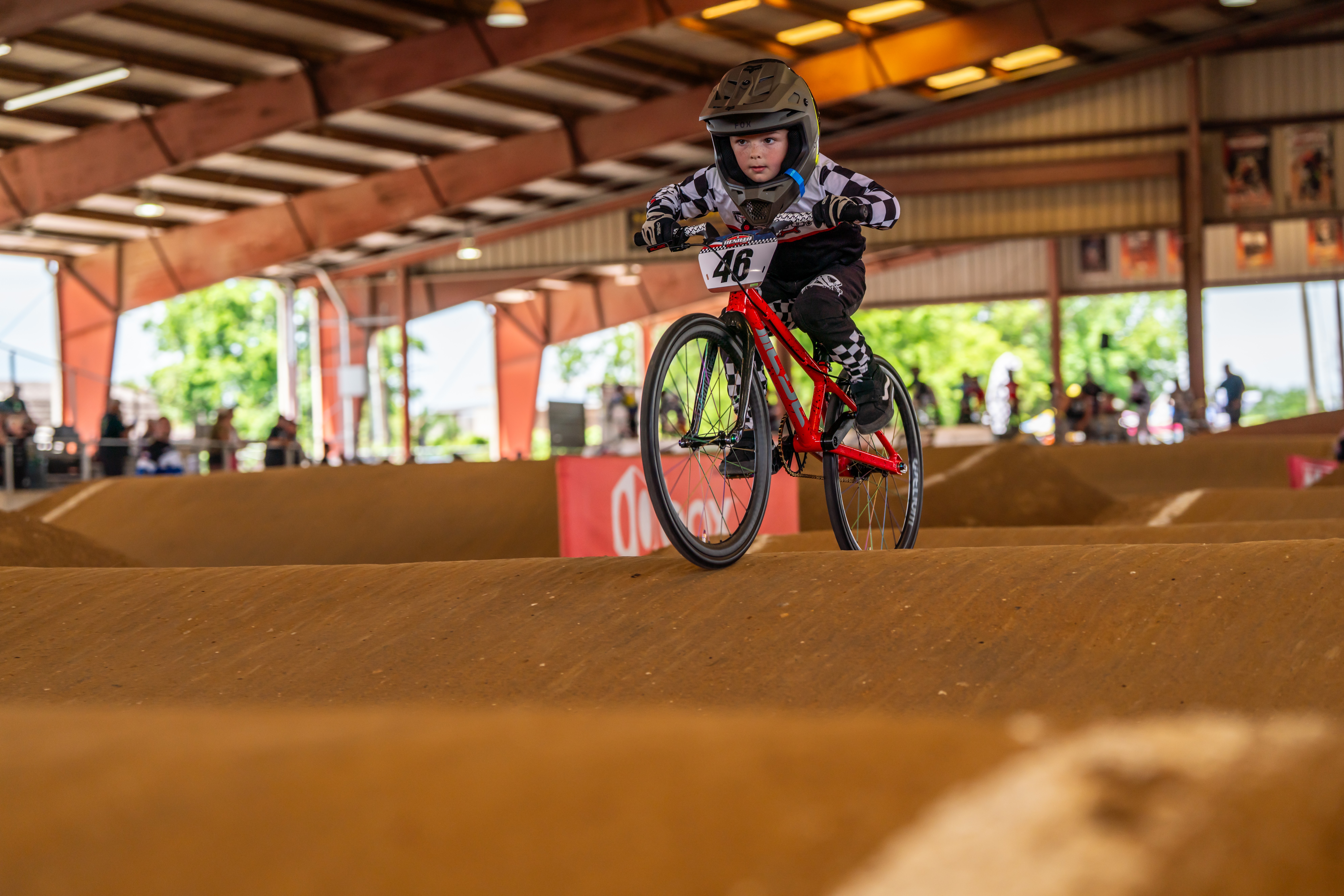 Child wearing helmet riding a bicycle on an indoor dirt track, with spectators and large windows in the background.