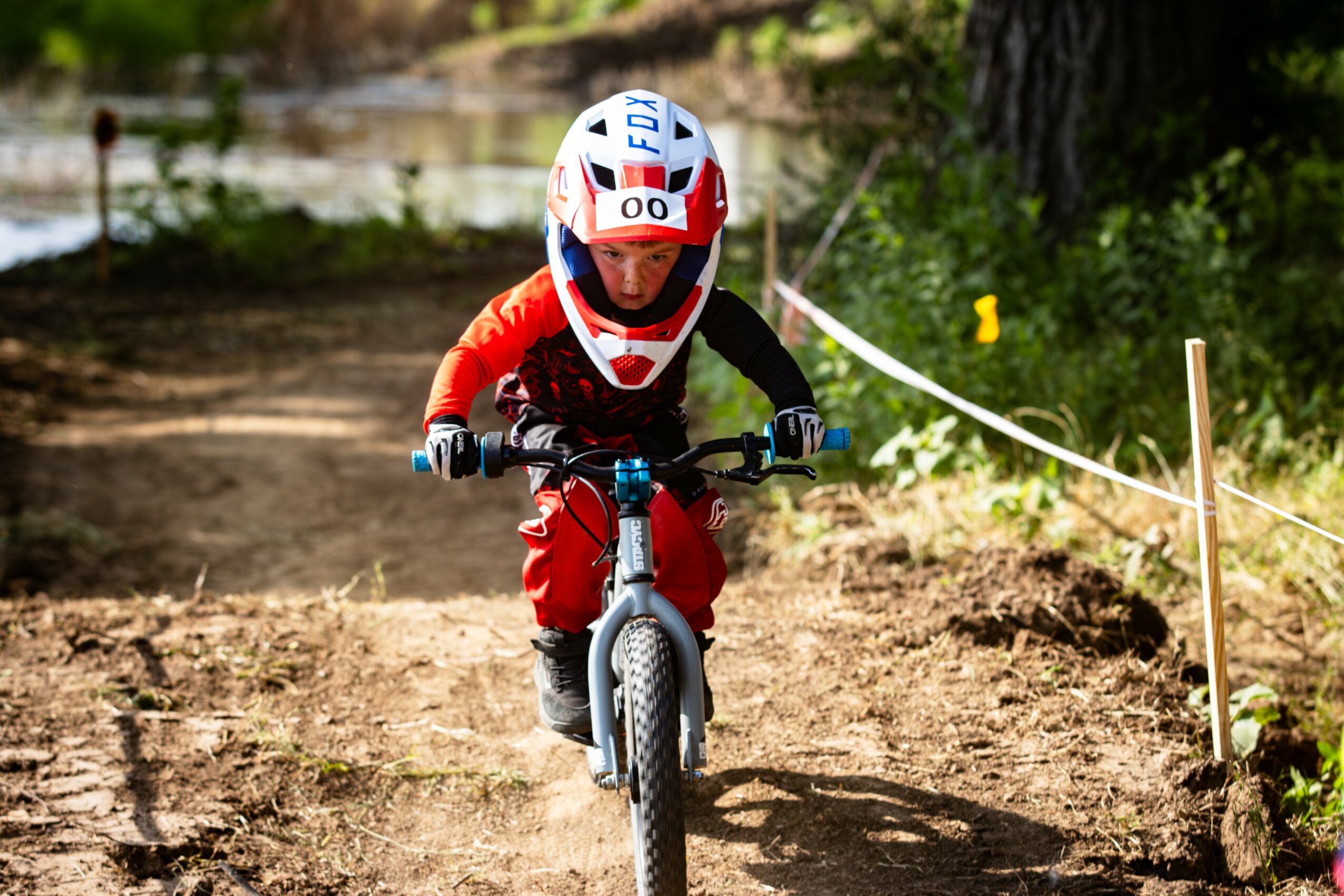 Child wearing a helmet and red outfit riding a mountain bike on a dirt trail in a forested area.