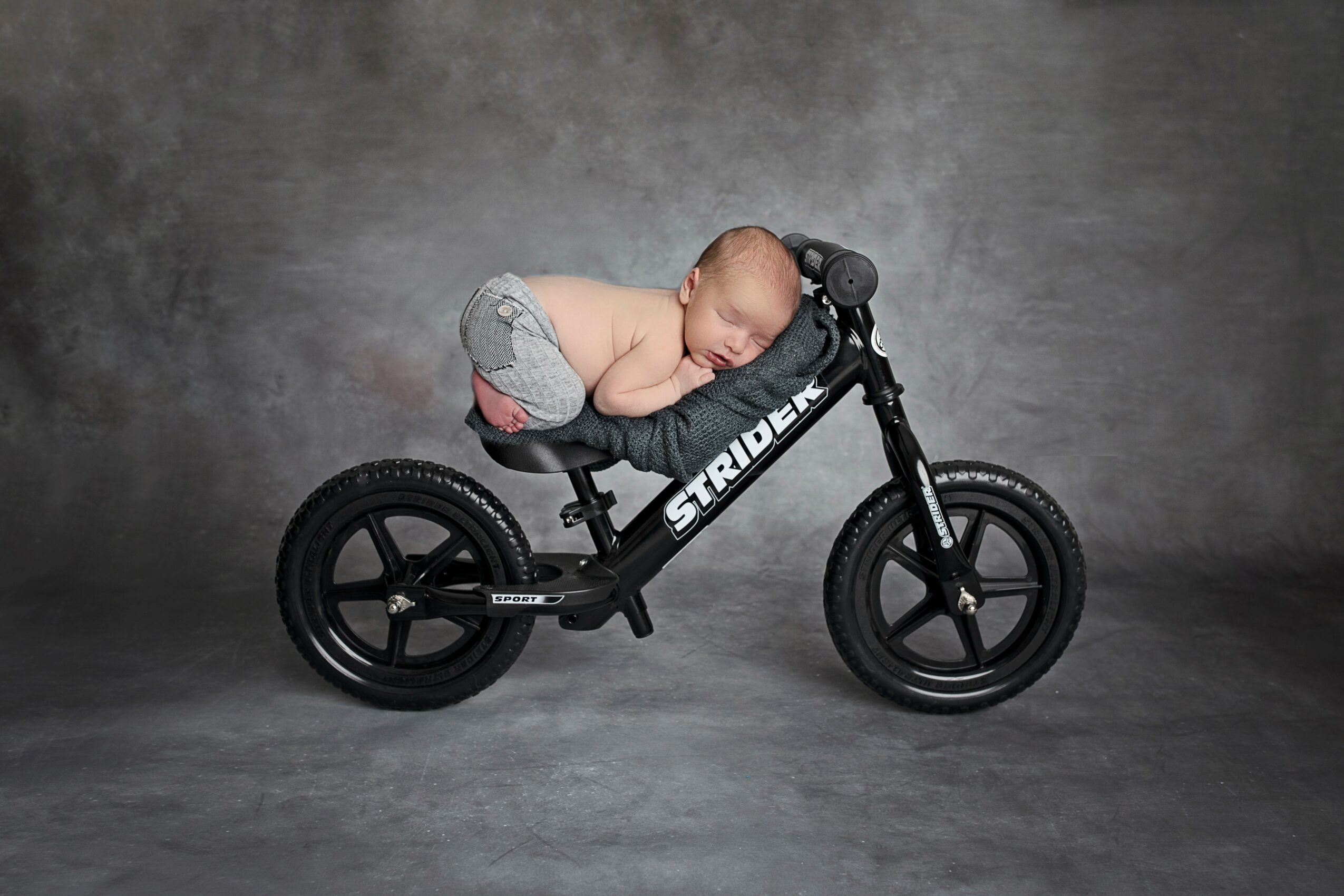 Baby sleeping on a small black balance bike with a gray background.