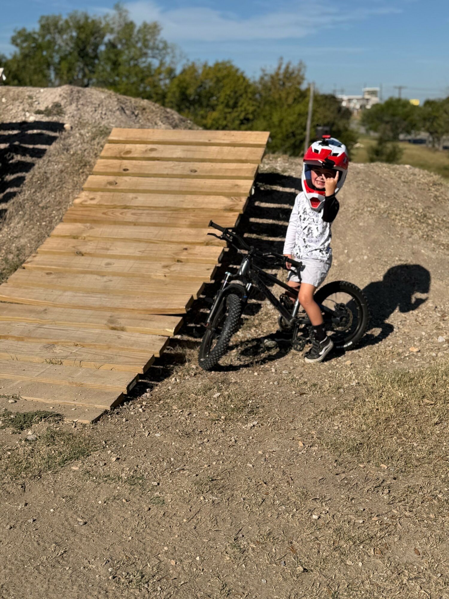 Child wearing a helmet and sunglasses standing next to a black mountain bike on a dirt ramp outdoors.