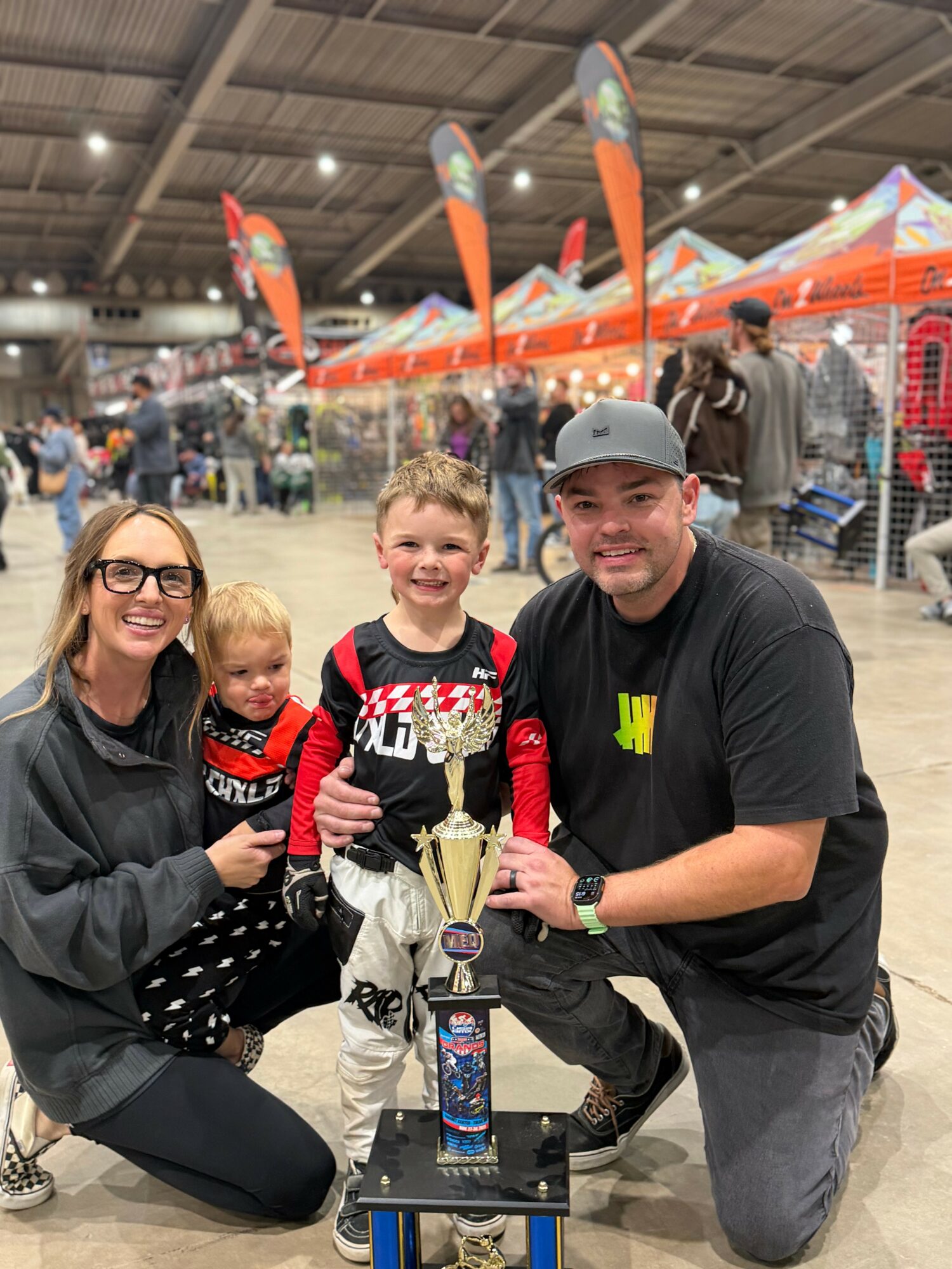 Family of four with a trophy at an indoor event, children smiling, adults kneeling, colorful tents in background.