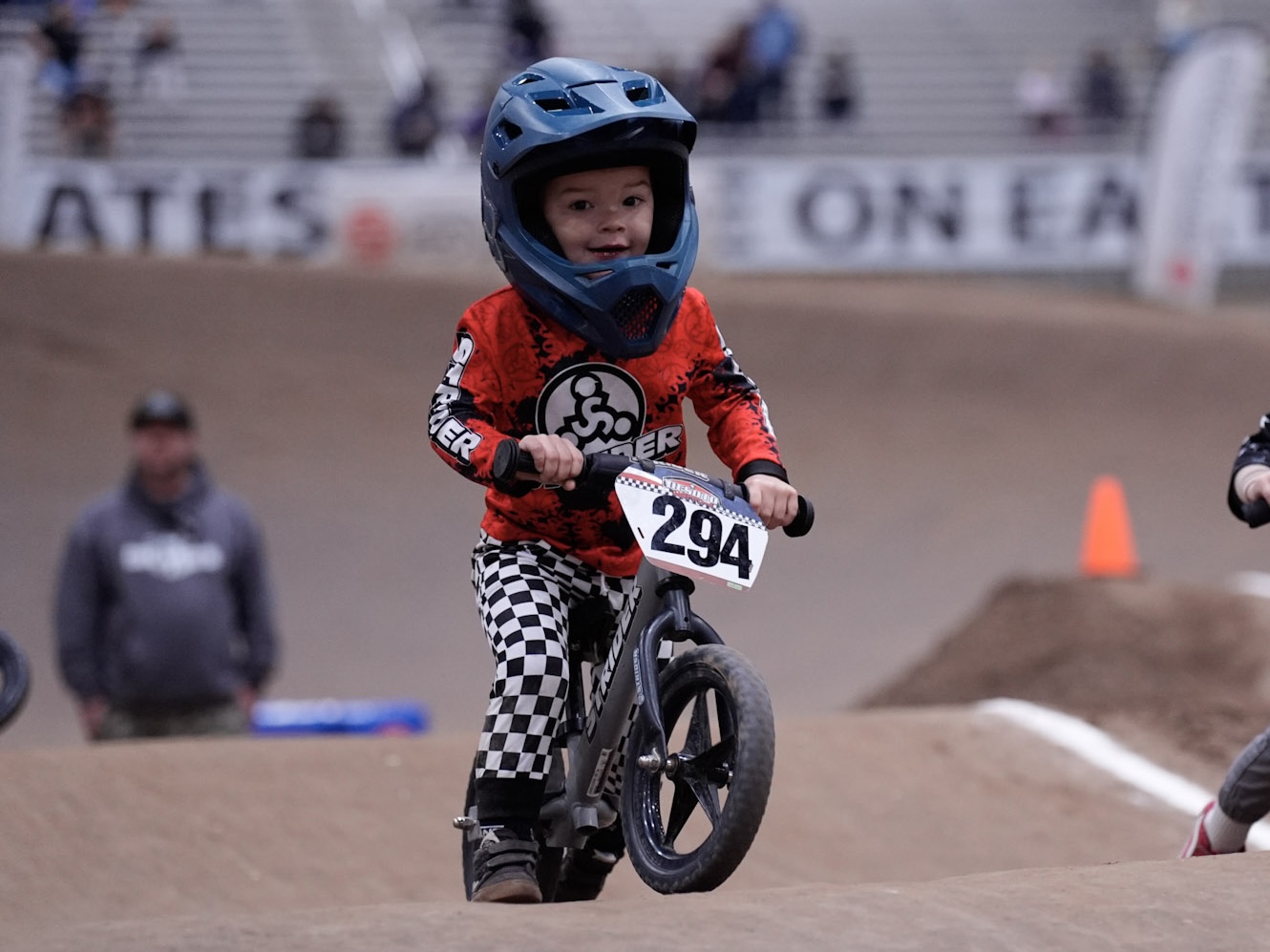 Child wearing a helmet and red jersey riding a bike on a dirt track, with spectators in the background.