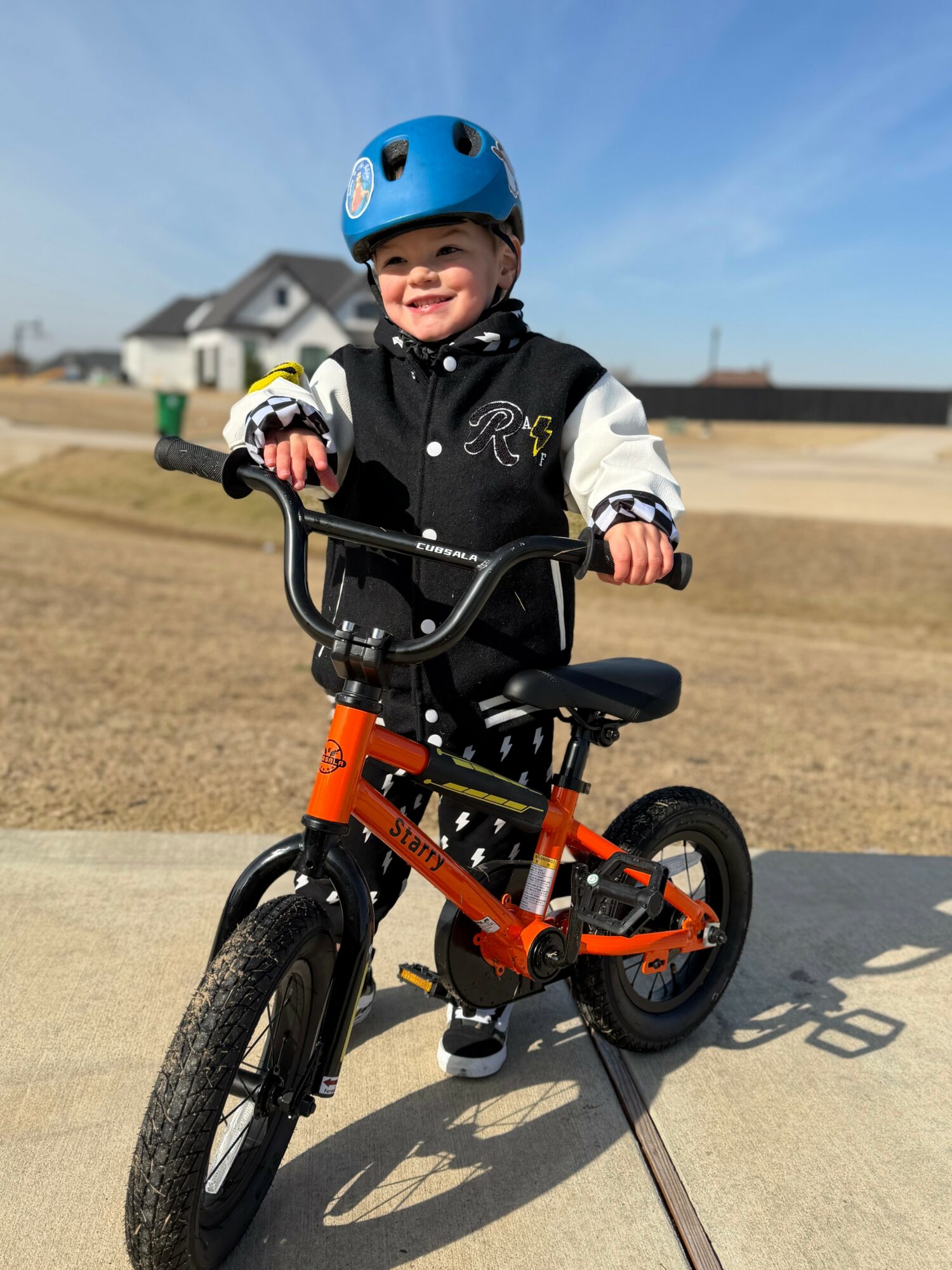 Child wearing a blue helmet and black jacket riding an orange bicycle outdoors on a sunny day.