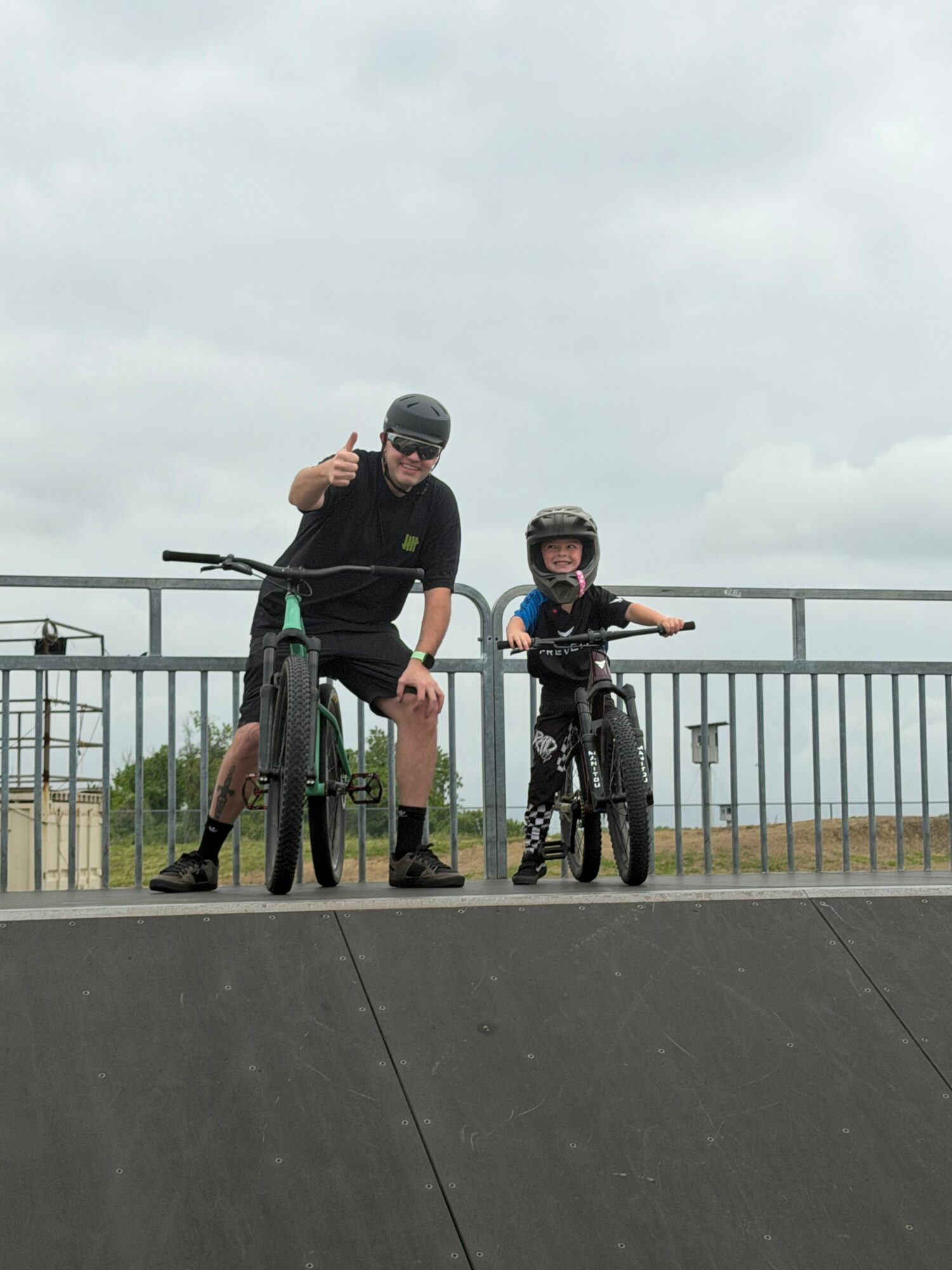 Two people on bicycles at a skate park, one giving a thumbs-up, overcast sky, metal railing, concrete surface.