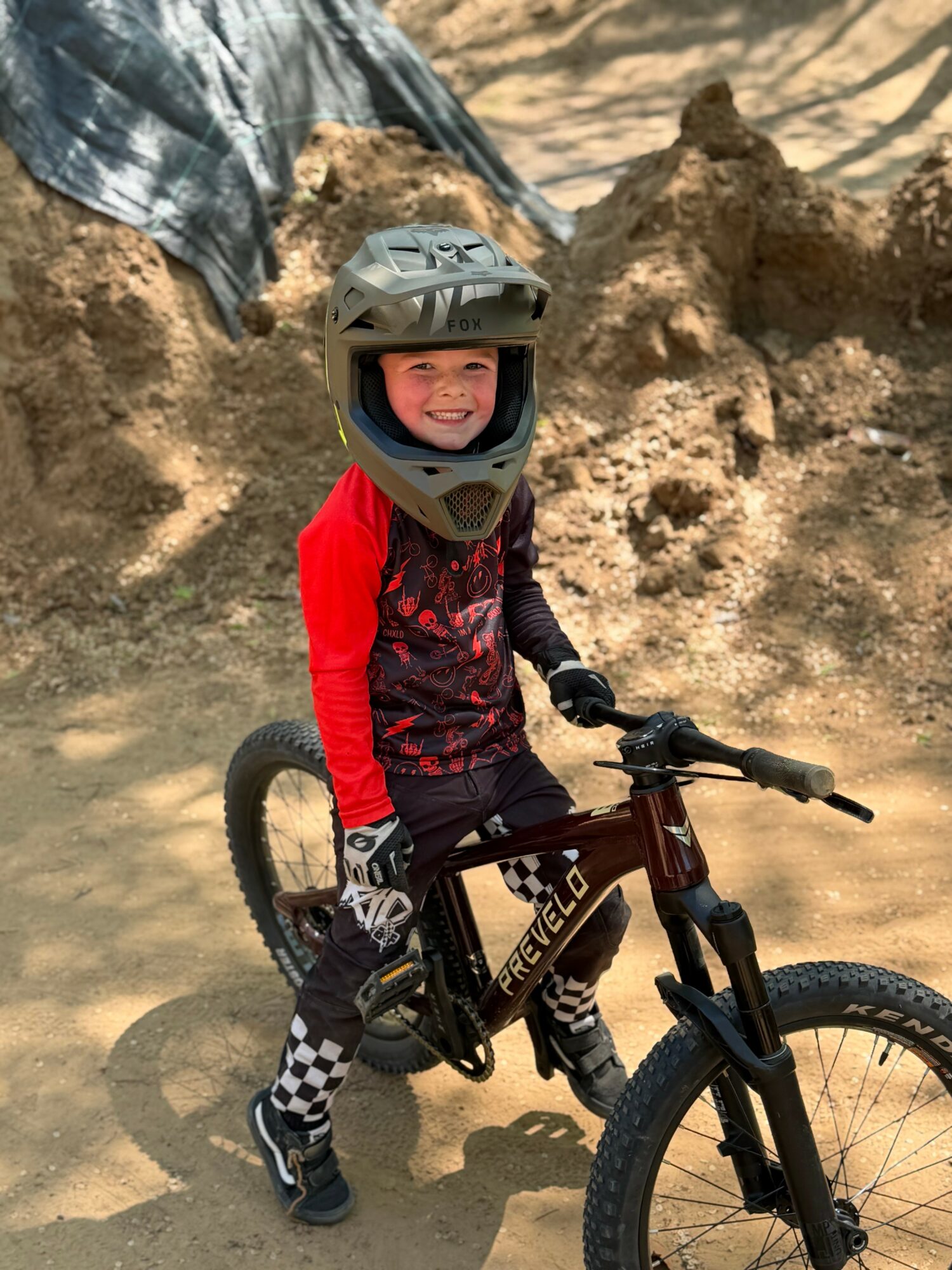 Child wearing a helmet and red and black cycling outfit, smiling on a mountain bike outdoors.