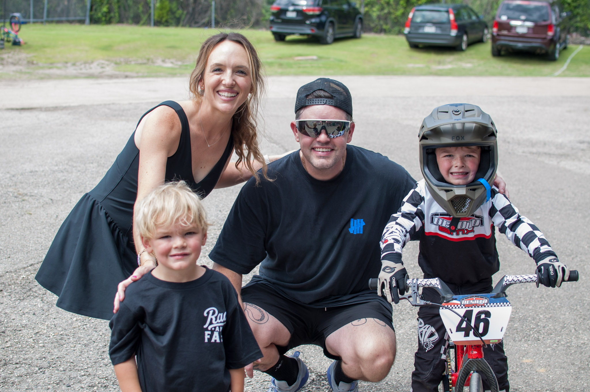 Four people, including a woman, two children, and a man, pose outdoors with a bike and cars in background.