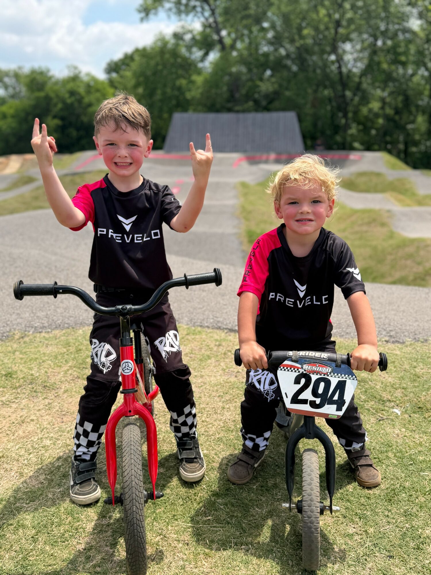 Two children with bikes outdoors, smiling, one making hand gestures, in a grassy area with trees and a skate park in background.