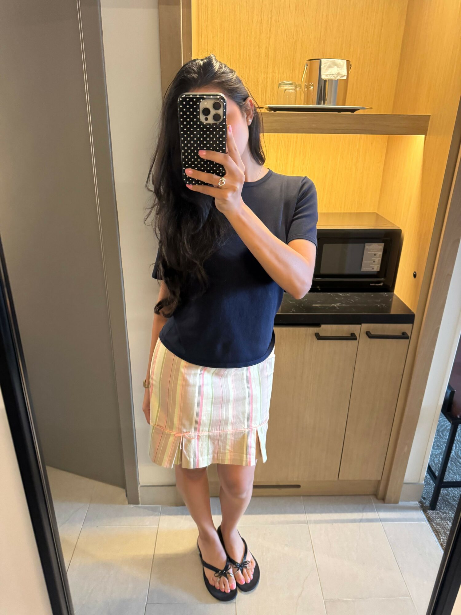 Person taking a mirror selfie in a kitchen, wearing a dark top, striped skirt, and flip-flops, holding a phone with a patterned case.