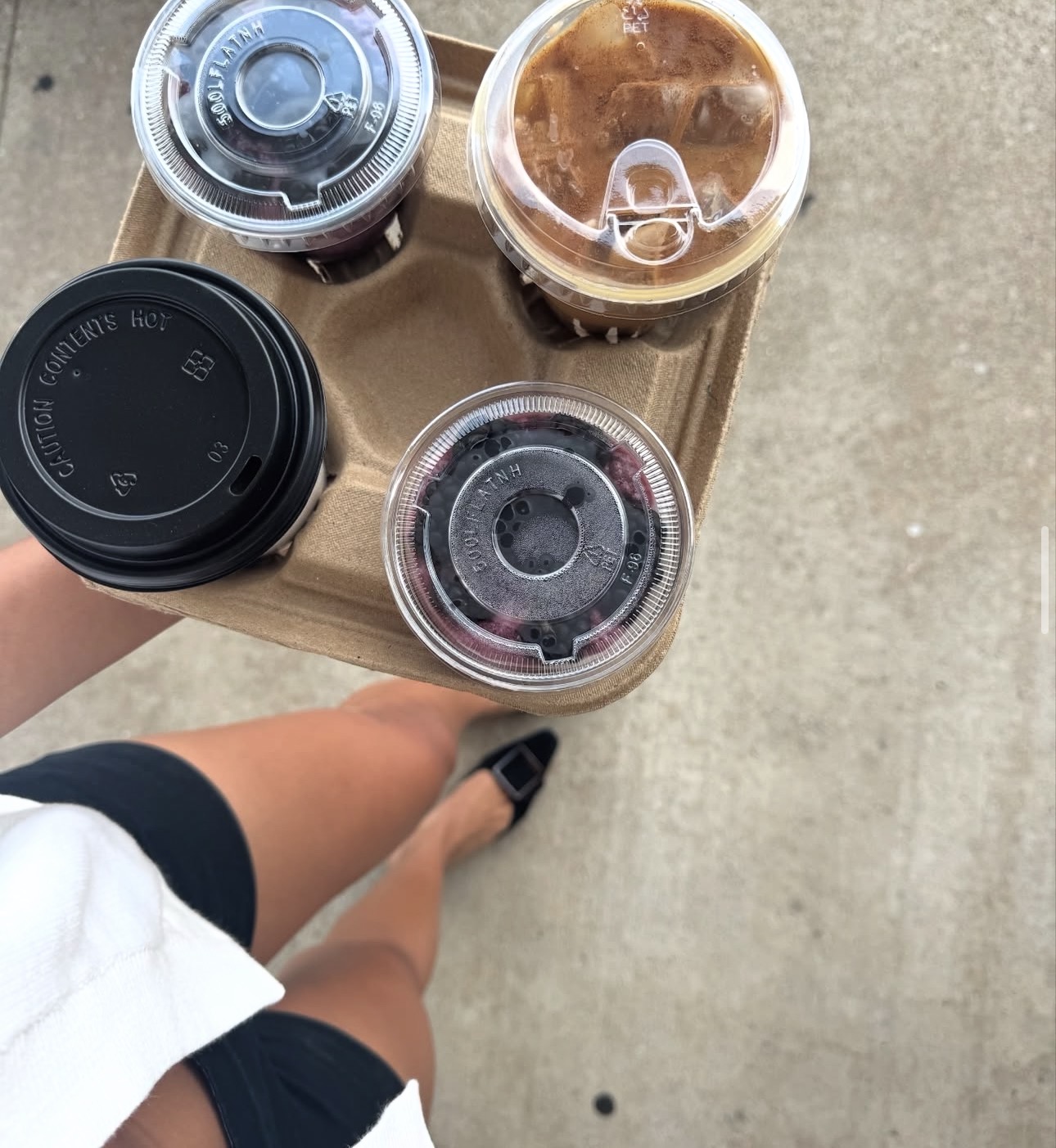 Person holding a tray with four cold drinks, two in plastic cups with lids, on a concrete surface.