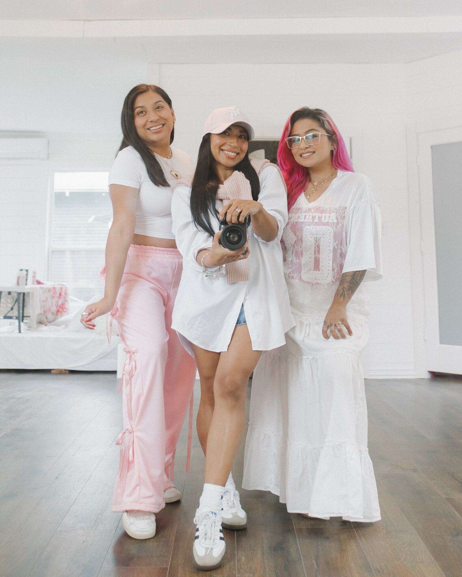 Three women standing together indoors, smiling, with one holding a camera, in a bright room with white walls and wooden floor.