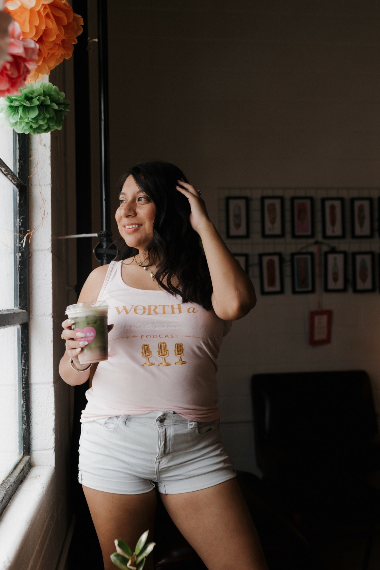 Woman with dark hair holding a drink, standing near a window with colorful paper flowers, smiling and looking outside.