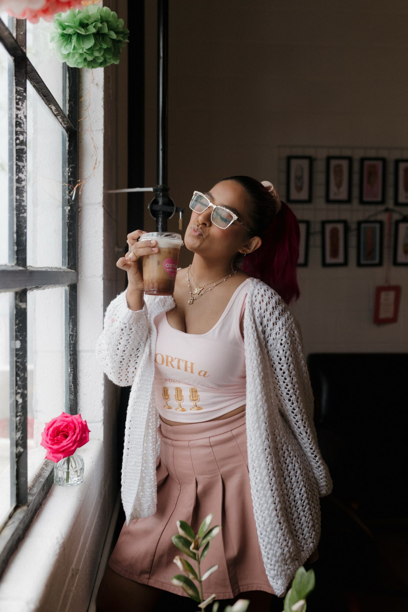 Woman with glasses drinks iced coffee near window, indoor setting with framed pictures, plants, and natural light.