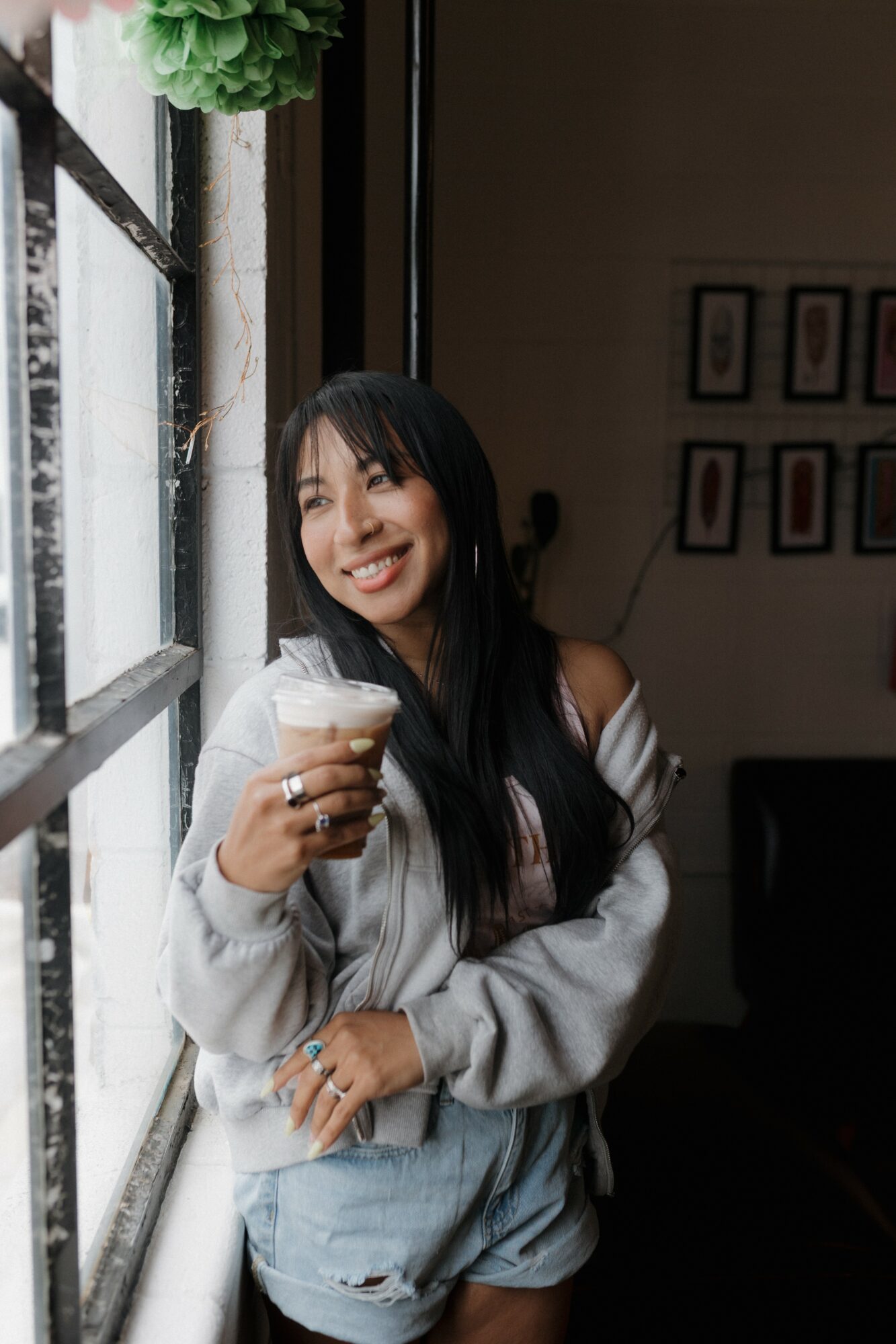 Young woman with long dark hair smiling, holding a drink, standing by a window in a cozy indoor setting.