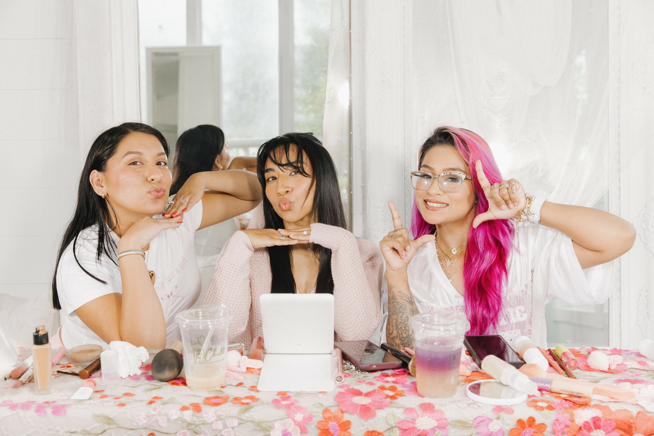 Three women sitting at a table with drinks and makeup, smiling and posing indoors with white curtains in background.