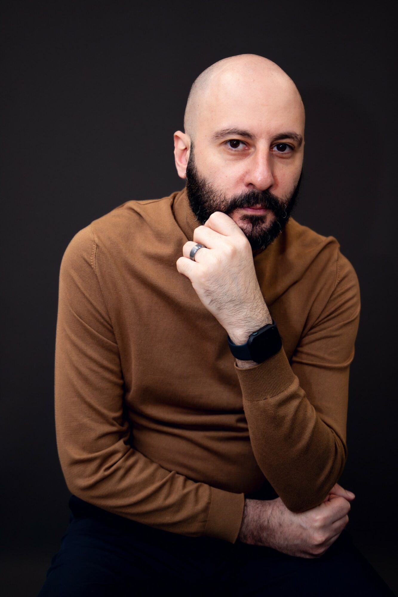 Bald man with beard wearing a brown long-sleeve shirt, resting chin on hand, against dark background.