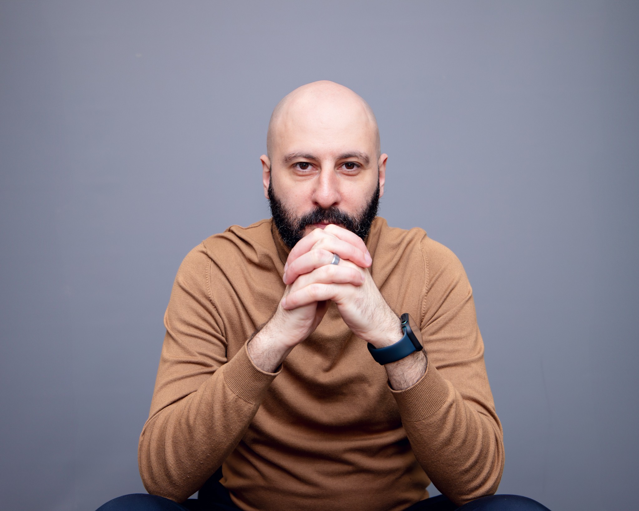 Bald man with a beard, wearing a brown long-sleeve shirt, sitting with hands clasped, looking at the camera, gray background.