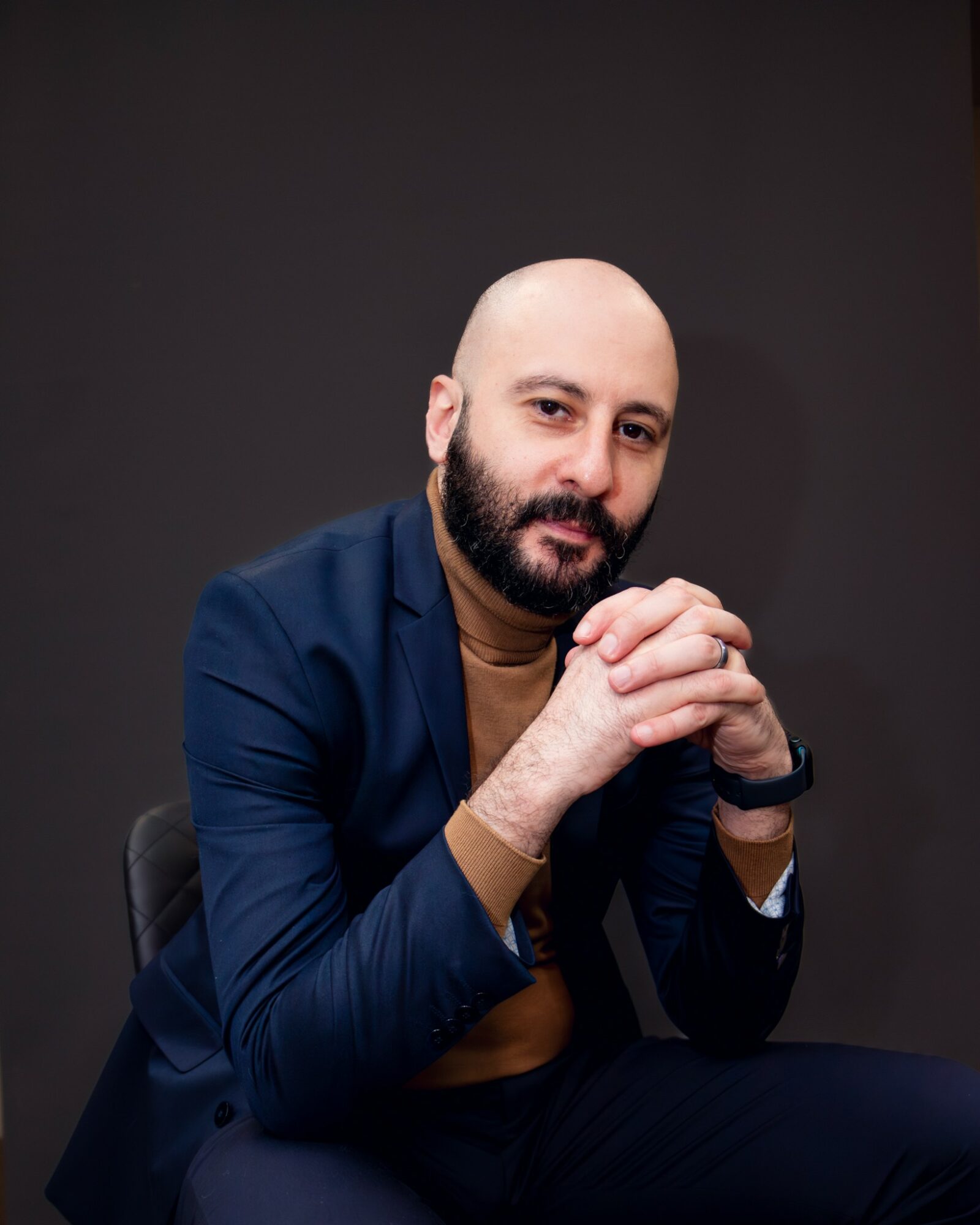 Man with a beard and bald head, wearing a navy blazer and brown shirt, sitting with hands clasped, against a dark background.
