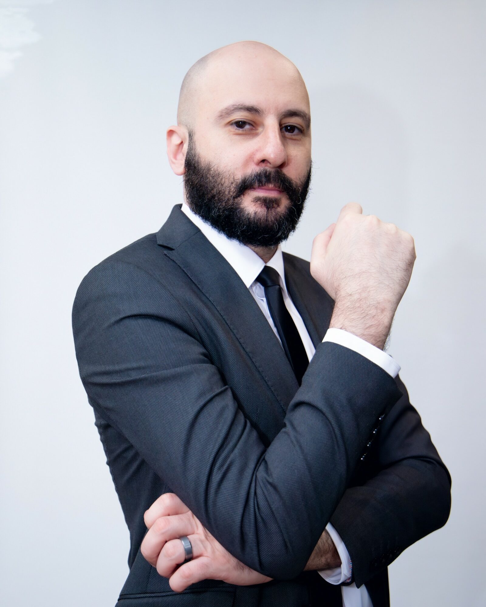 Bald man with beard in a suit, crossing arms, looking confident, against a plain light background.