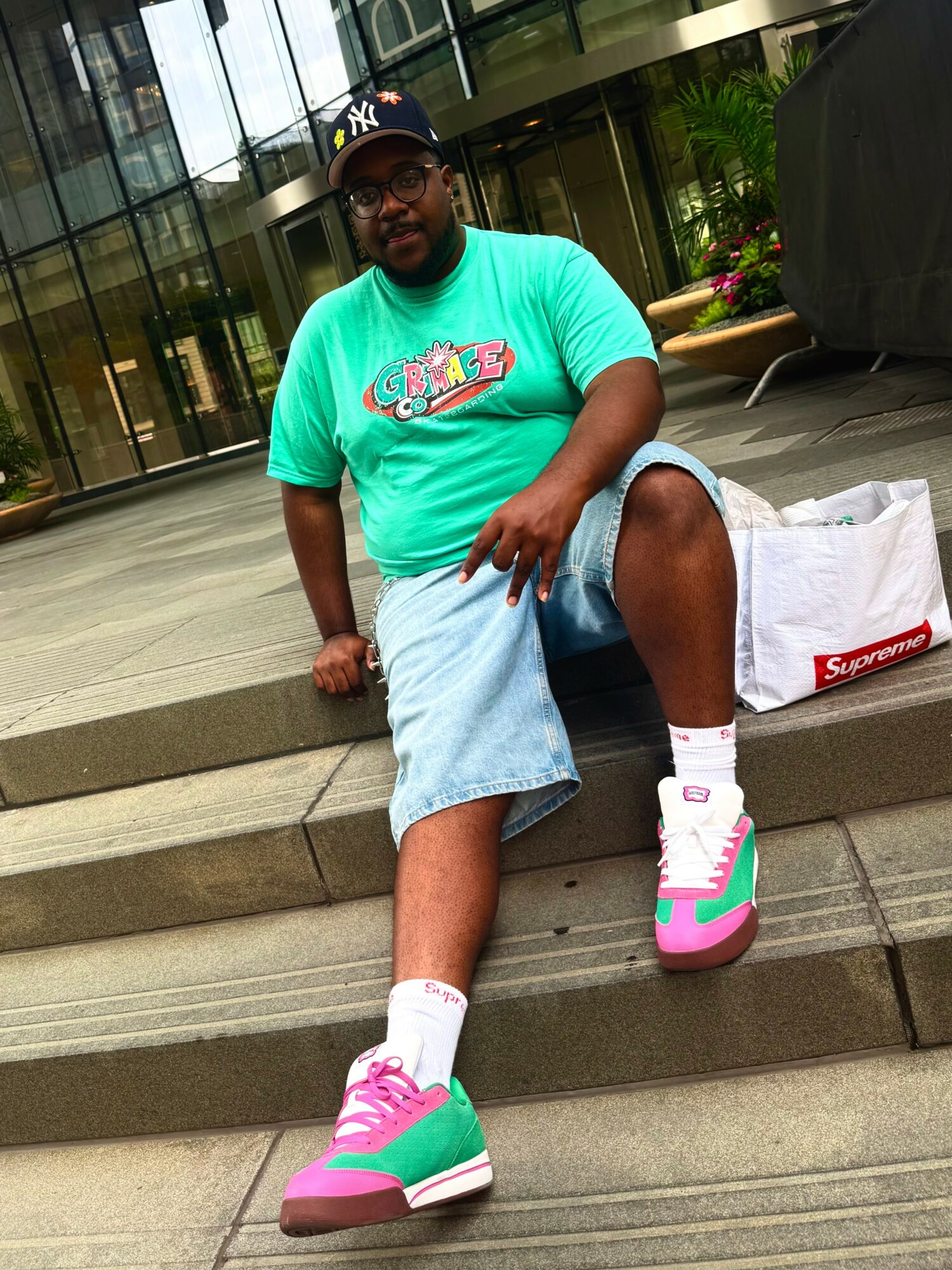 Person sitting on steps outdoors, wearing a turquoise T-shirt, denim shorts, pink and teal sneakers, and a black cap.