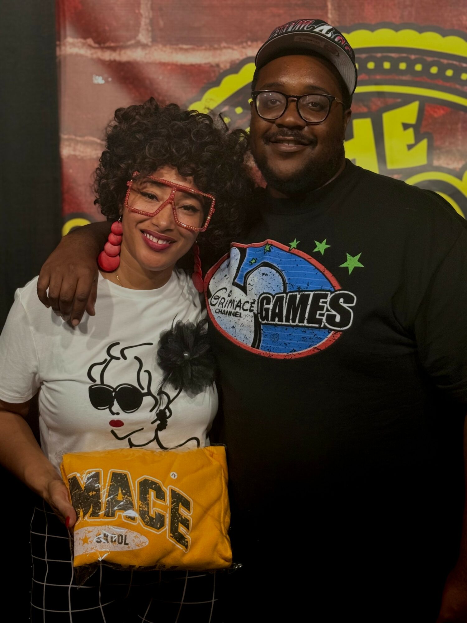 Two people smiling, one holding a yellow bag, standing in front of a colorful background with a logo.