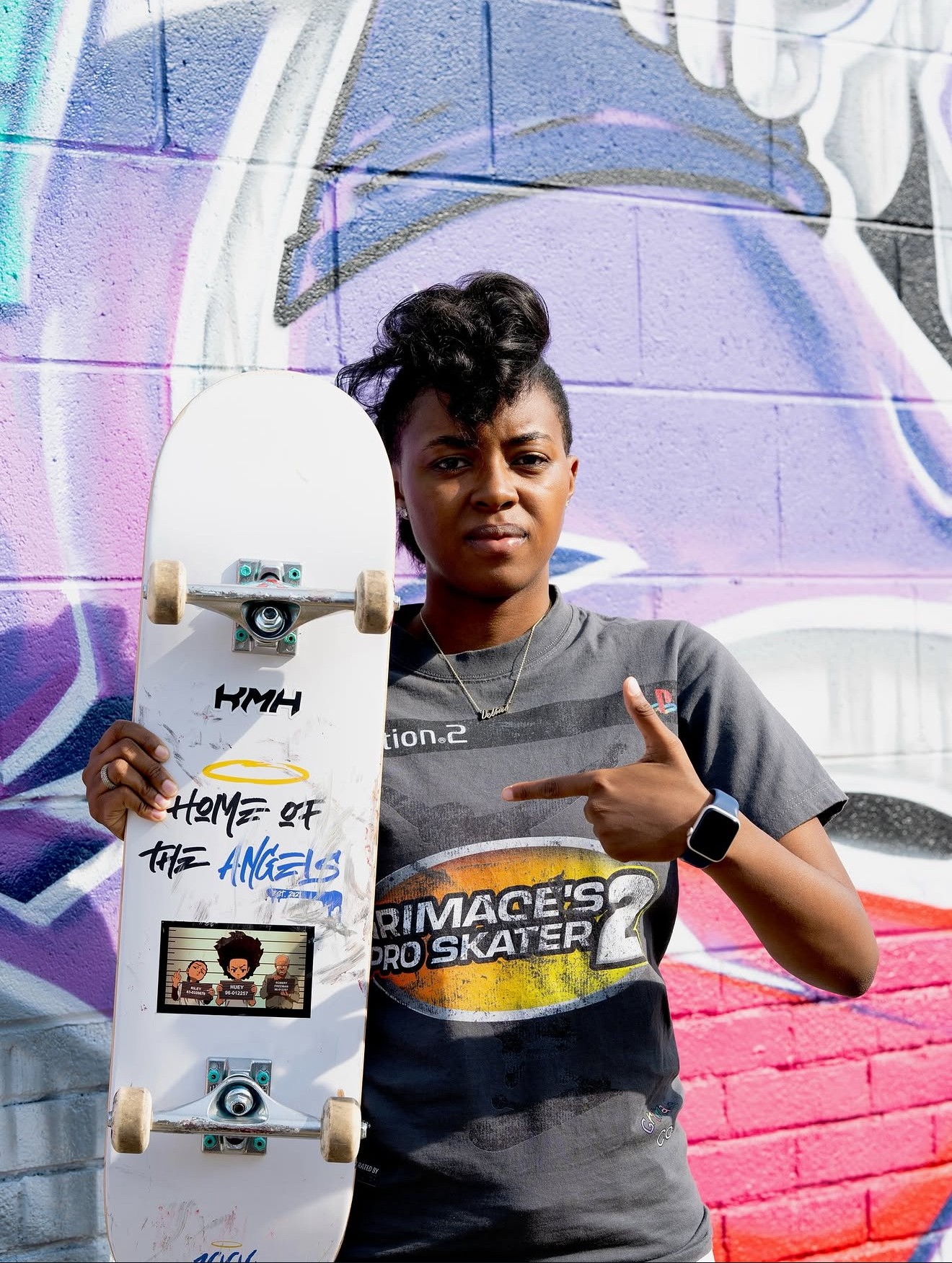 Young woman holding skateboard in front of colorful graffiti wall, pointing at skateboard, wearing a gray T-shirt.