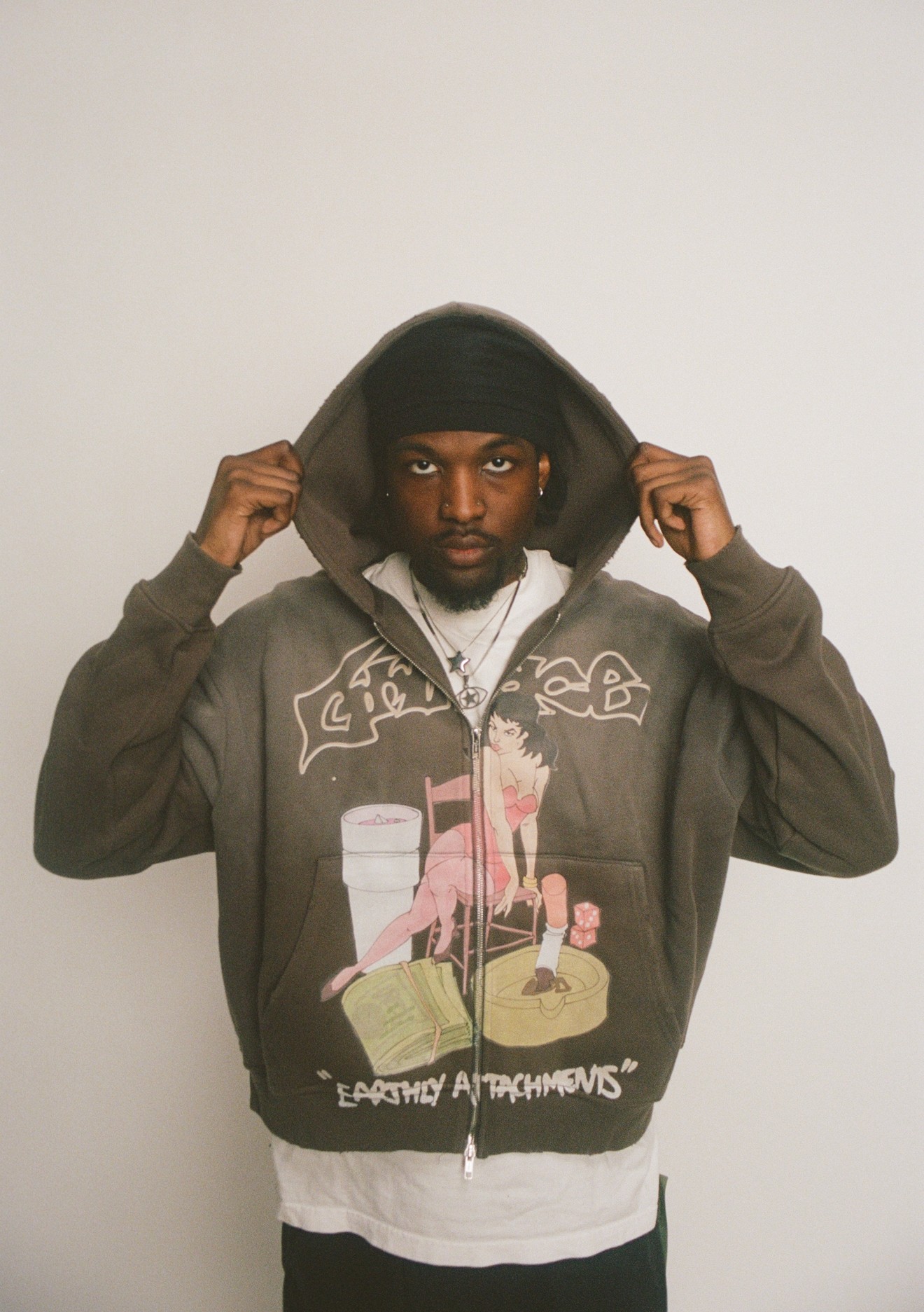 Man with dark hoodie and jacket, holding the hood with both hands, facing forward, against a plain background.