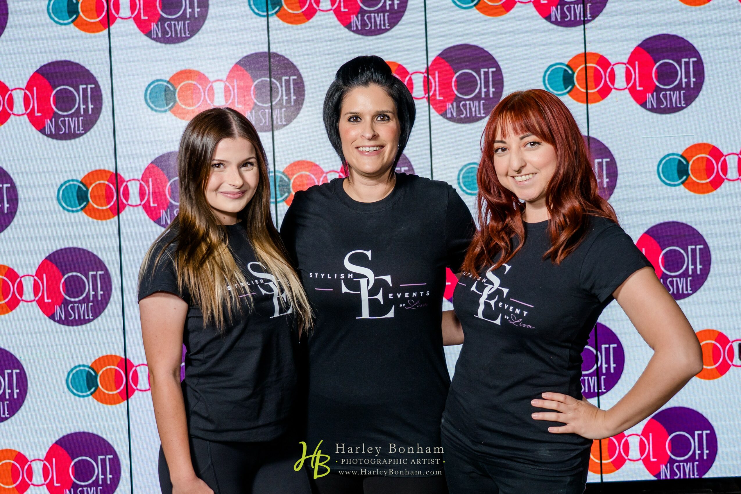 Three women standing together in front of a colorful backdrop with logos, smiling at the camera.