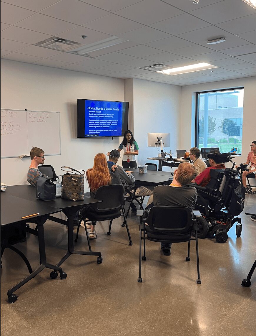 A woman presents to a group in a classroom with a large screen displaying a blue slide, whiteboard, and window.