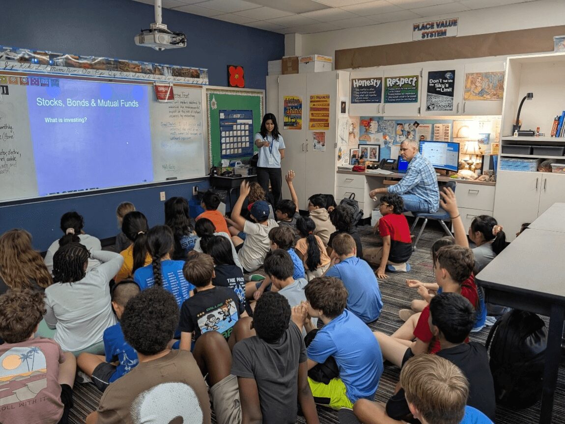 Teacher standing at front of classroom with students seated on floor, presentation screen displaying financial topics, classroom with educational posters.