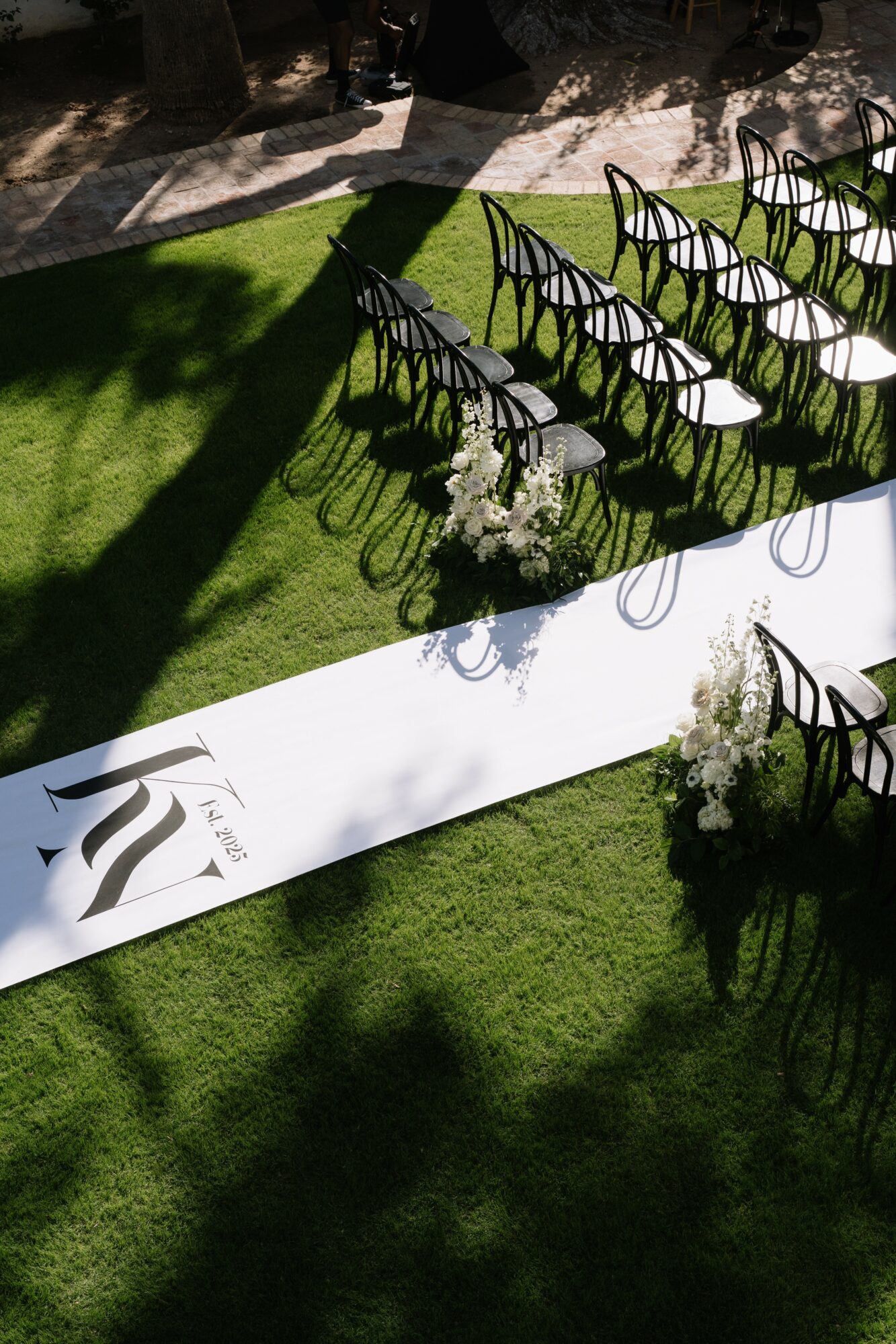 Outdoor event setup with chairs arranged on grass, shadowed area, and a white aisle runner with a logo, flowers at the end.