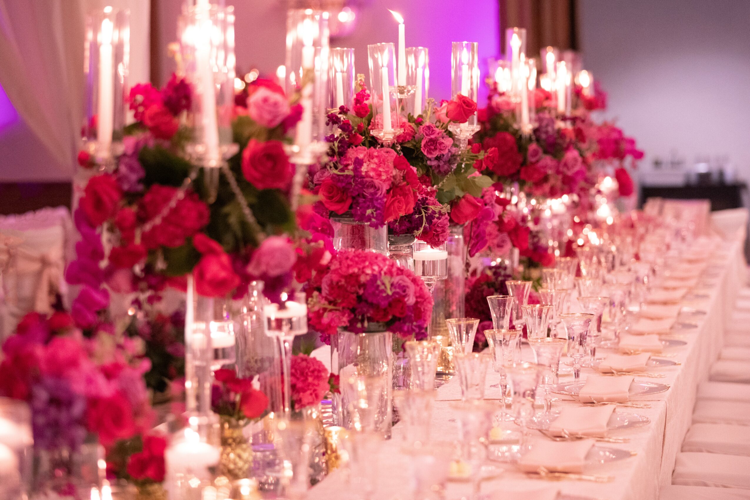 Long table decorated with pink and purple flowers in tall glass vases, surrounded by lit candles, set for an event.